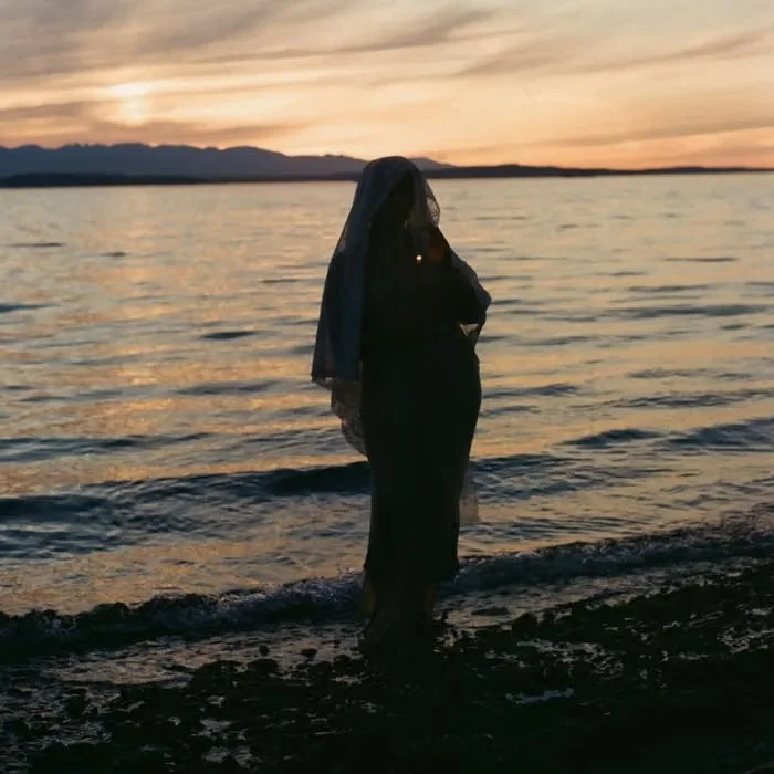 A woman standing in the water at sunset, wearing a dress and a veil over her head, with mountains and a colorful sky in the background.