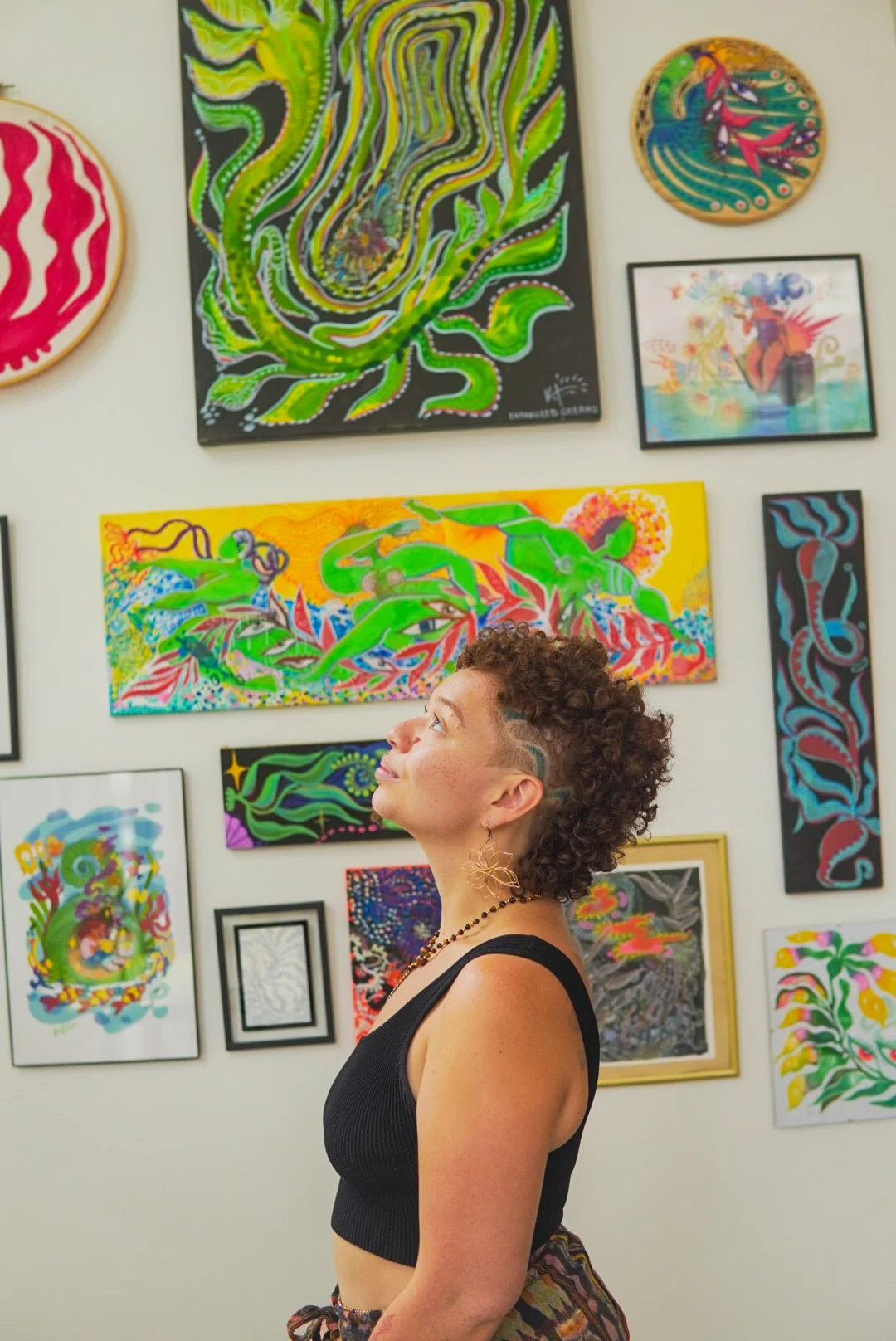 Woman with curly hair, wearing a black sleeveless top, and gold earrings, standing in front of colorful abstract artwork in a gallery.
