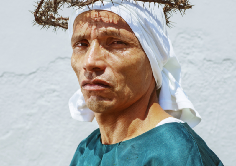 Portrait of a man with a serious expression, wearing a white head covering and a crown-like headpiece made of branches, standing against a white brick wall.