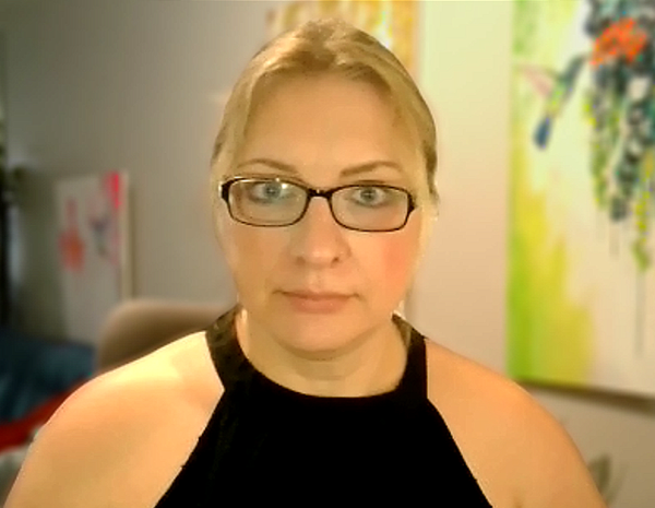 A woman with glasses and blonde hair, wearing a black sleeveless top, looking softly at the camera in an indoor setting.