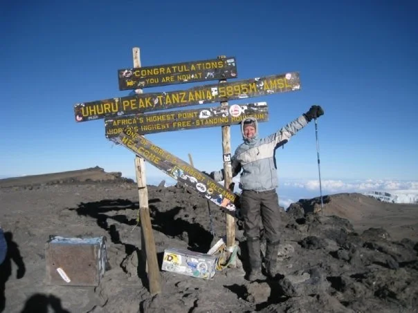 Person standing next to a signpost at Uhuru Peak on Mount Kilimanjaro, Tanzania, with clear skies in the background. The sign indicates the summit's elevation of 5895 meters.