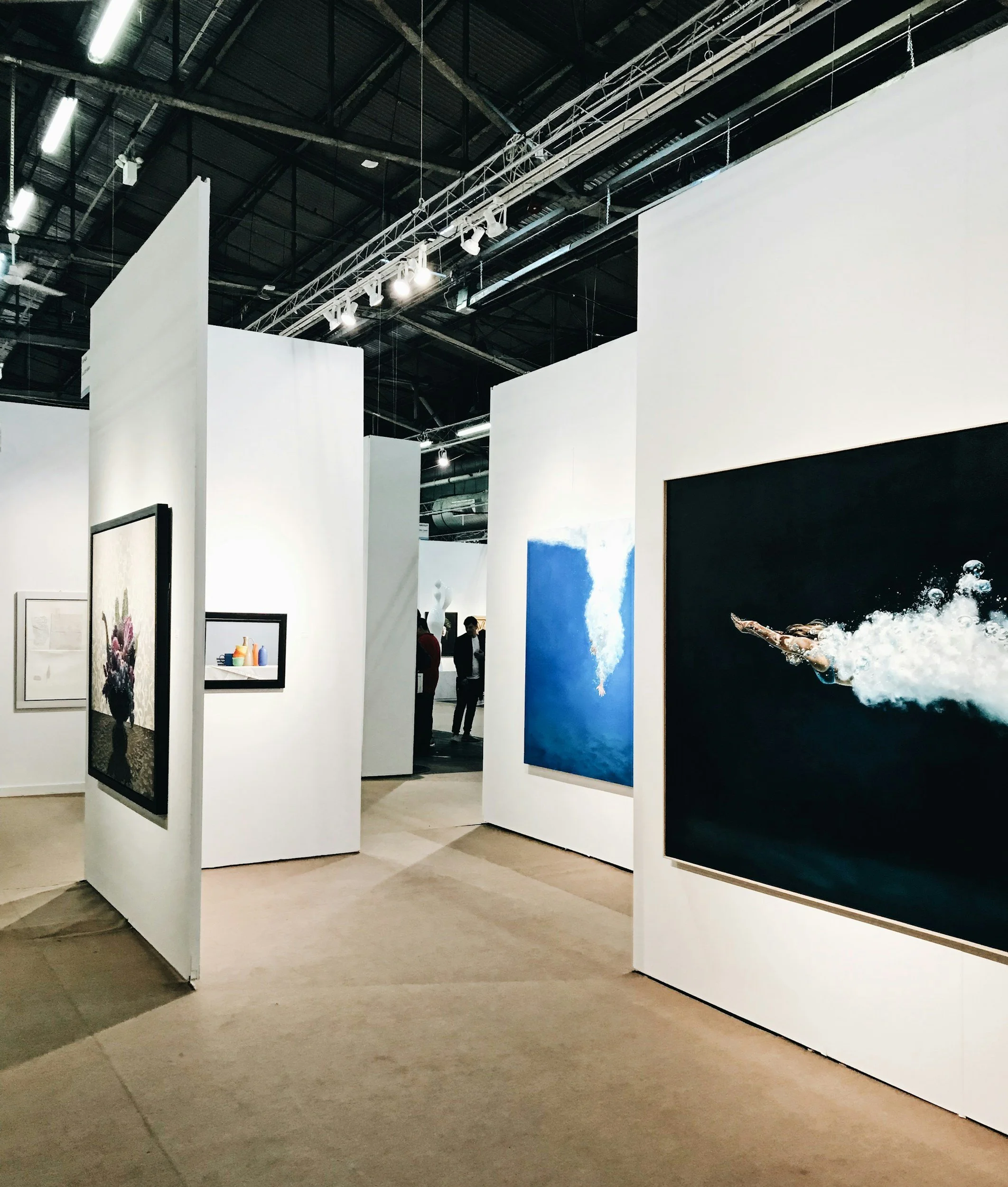 Interior of an art gallery with white partition walls displaying various artworks, including photographs and paintings, under industrial ceiling lighting.
