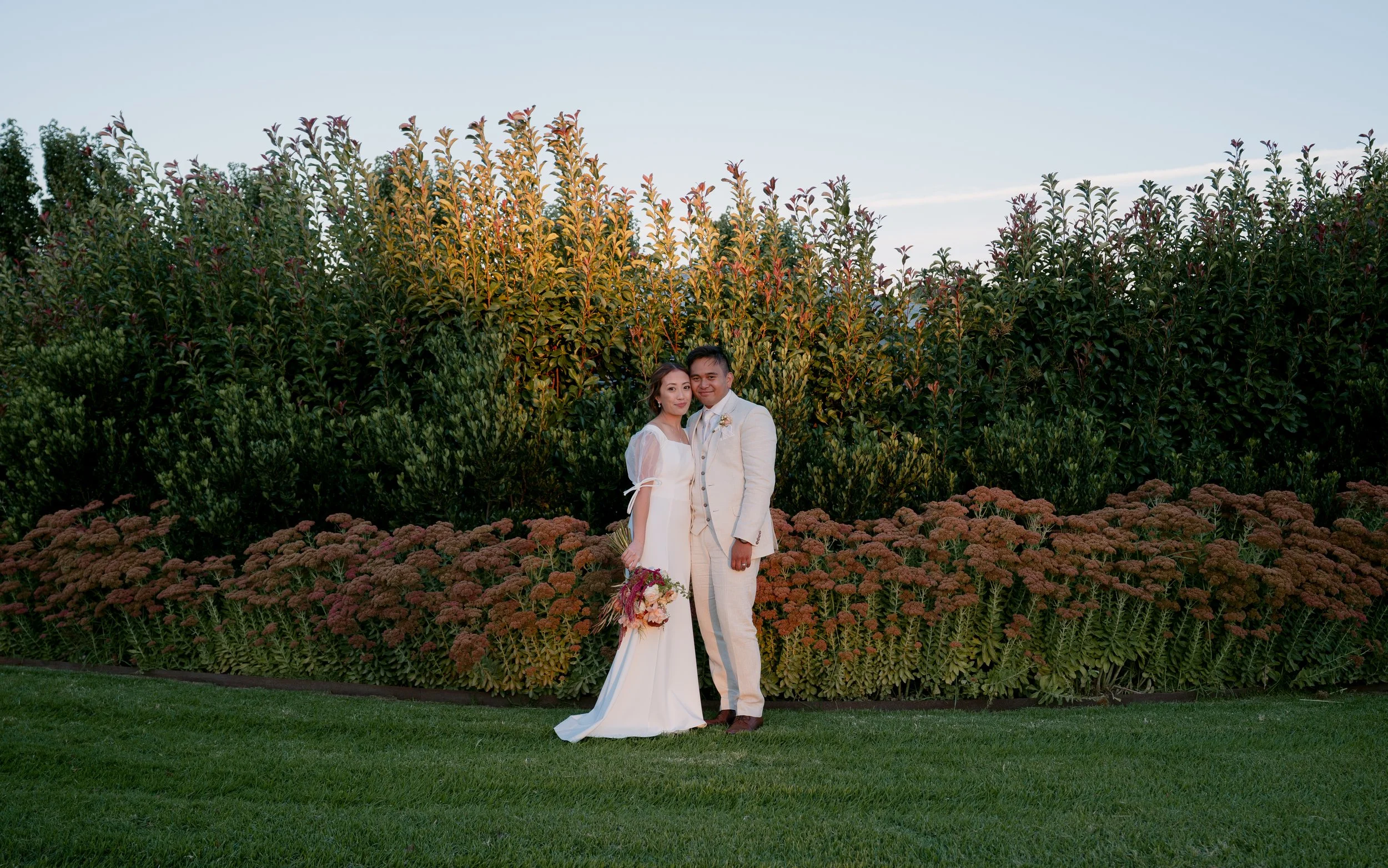 A bride and groom stand together outdoors on a grassy area at Olivehouse at Greendale Grove with trees and shrubs behind them, celebrating their wedding.