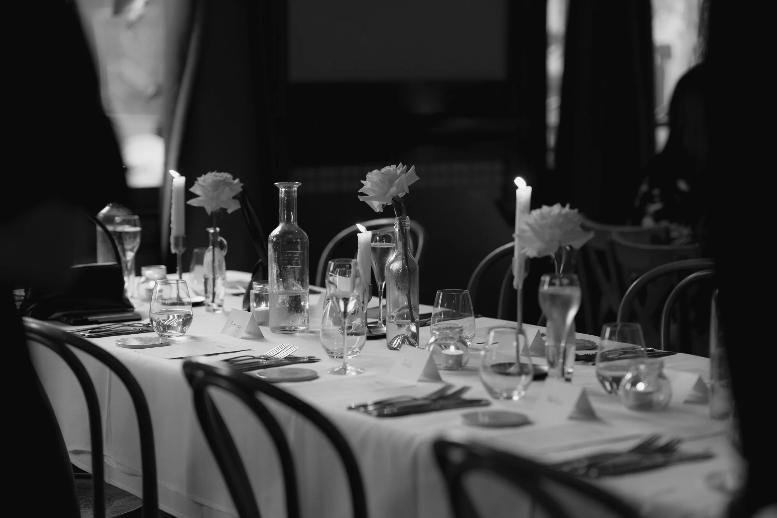 Table set with glasses, cutlery, vases with flowers and lit candles in a dimly lit room, black and white photograph.