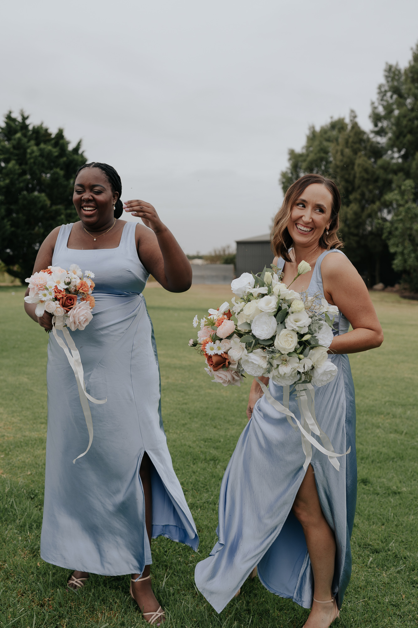 Two women in matching light blue dresses holding bouquets of white, peach, and pink flowers, standing on a grassy field with trees and a building behind them, smiling and laughing. Witchmount Estate Wedding