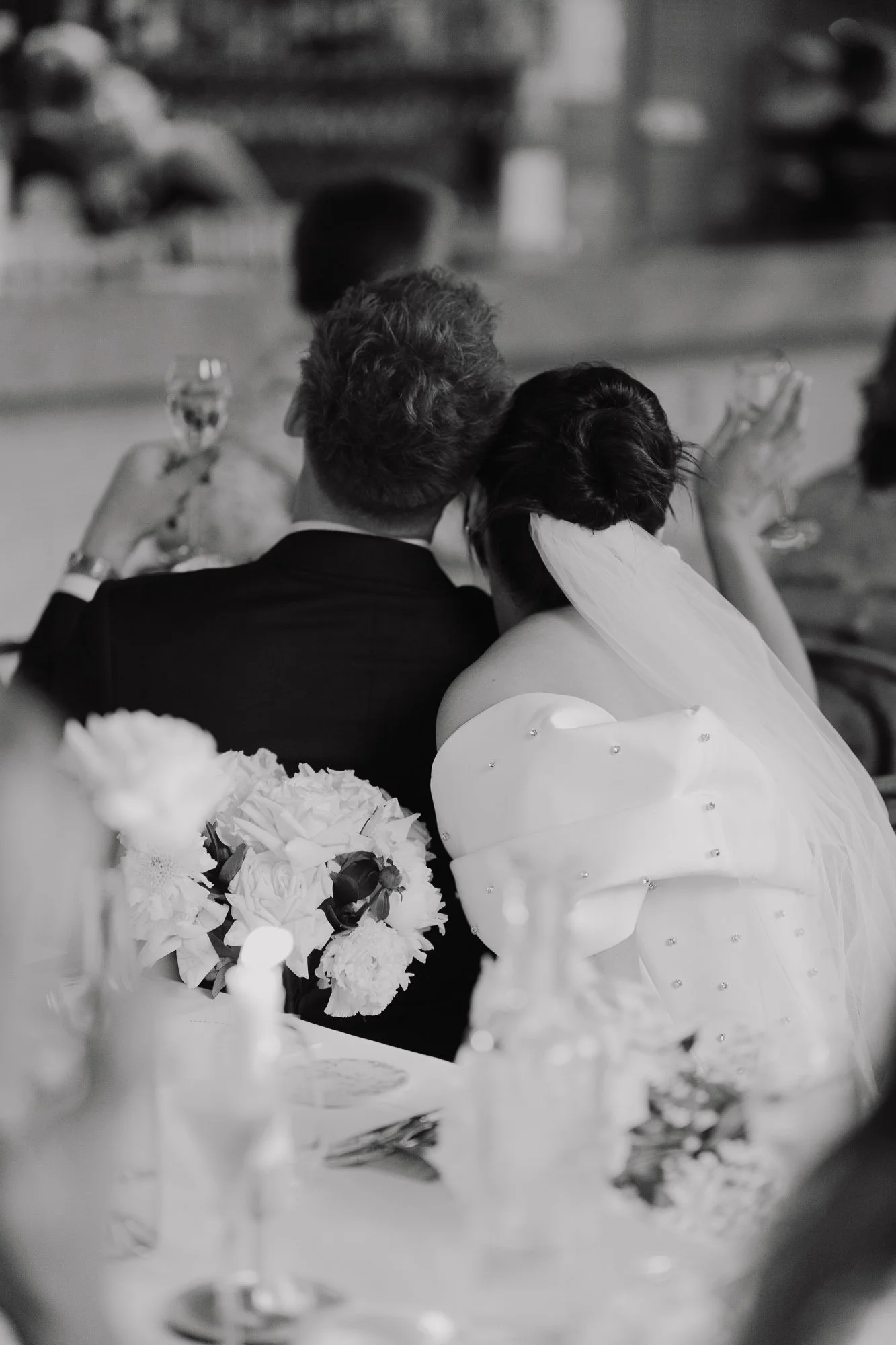 Black and white photo of a bride and groom at a wedding reception, sitting closely with the bride resting her head on the groom's shoulder. The bride is wearing a wedding dress with an off-the-shoulder design and veil, and holding a bouquet of flower