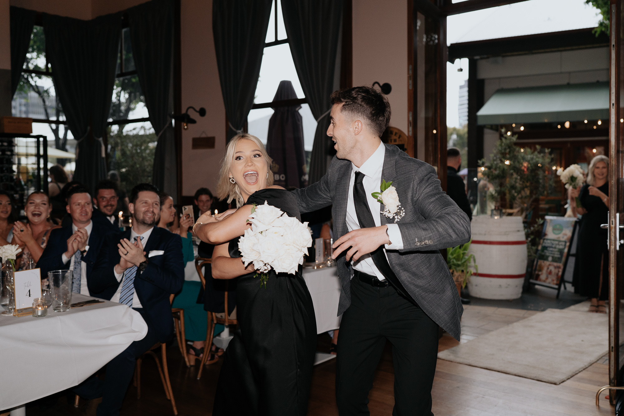A wedding reception with a woman in a black dress and a man in a gray suit dancing and laughing, while guests sit at tables clapping and smiling in the background.