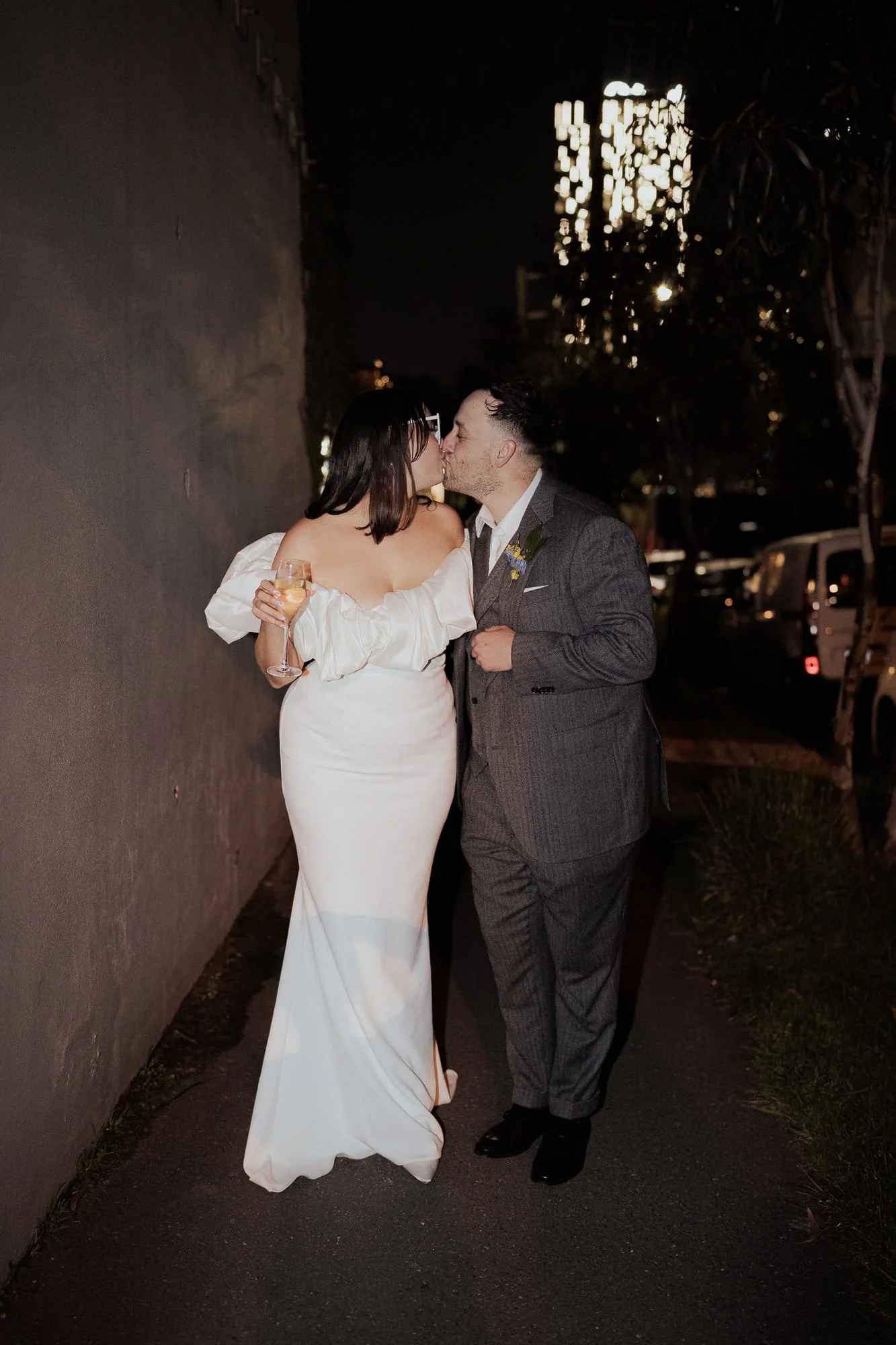 Melbourne Wedding Photography. A newlywed couple sharing a kiss at night outside Half Acre, a food&desire venue in South Melbourne, with the woman holding a glass of champagne, in front of a dark wall and parked cars.