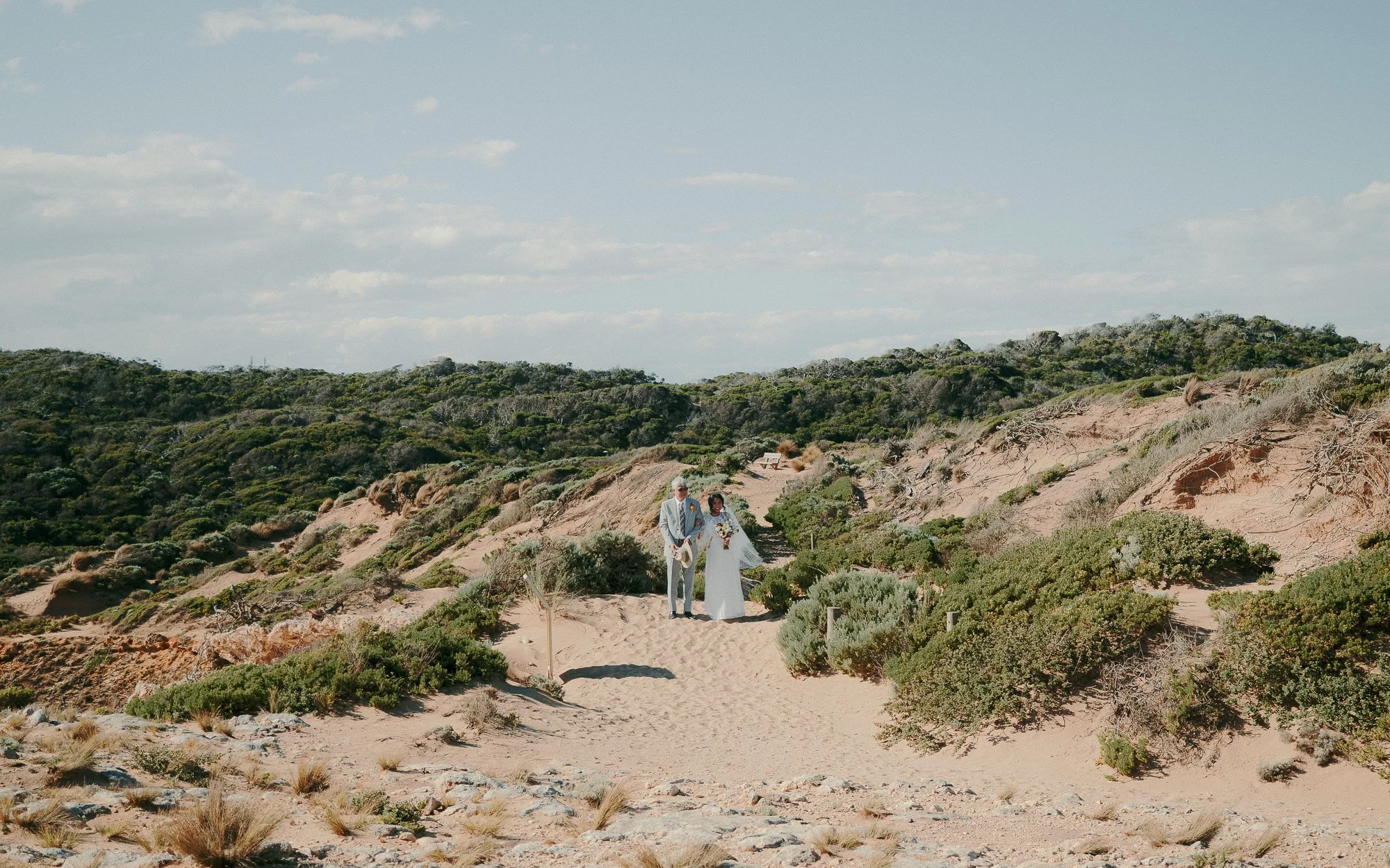 Bride and groom walking on a sandy path through a desert landscape with bushes, wearing wedding attire.