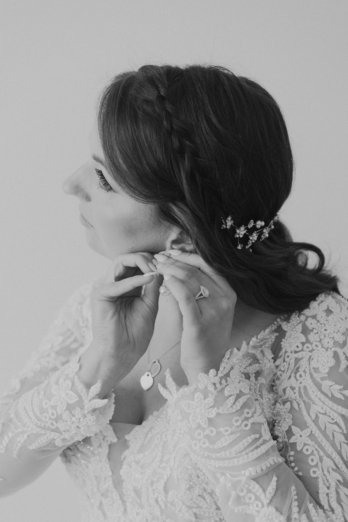 A bride with dark hair styled in loose waves, wearing a lace wedding dress, putting on earrings with one hand, displaying a ring on her finger, and wearing jewelry including a hair accessory and necklace.