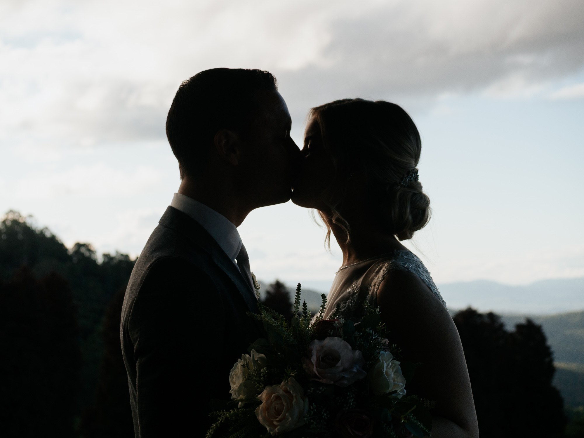 Silhouette of a bride and groom kissing outside with mountains and trees in the background.