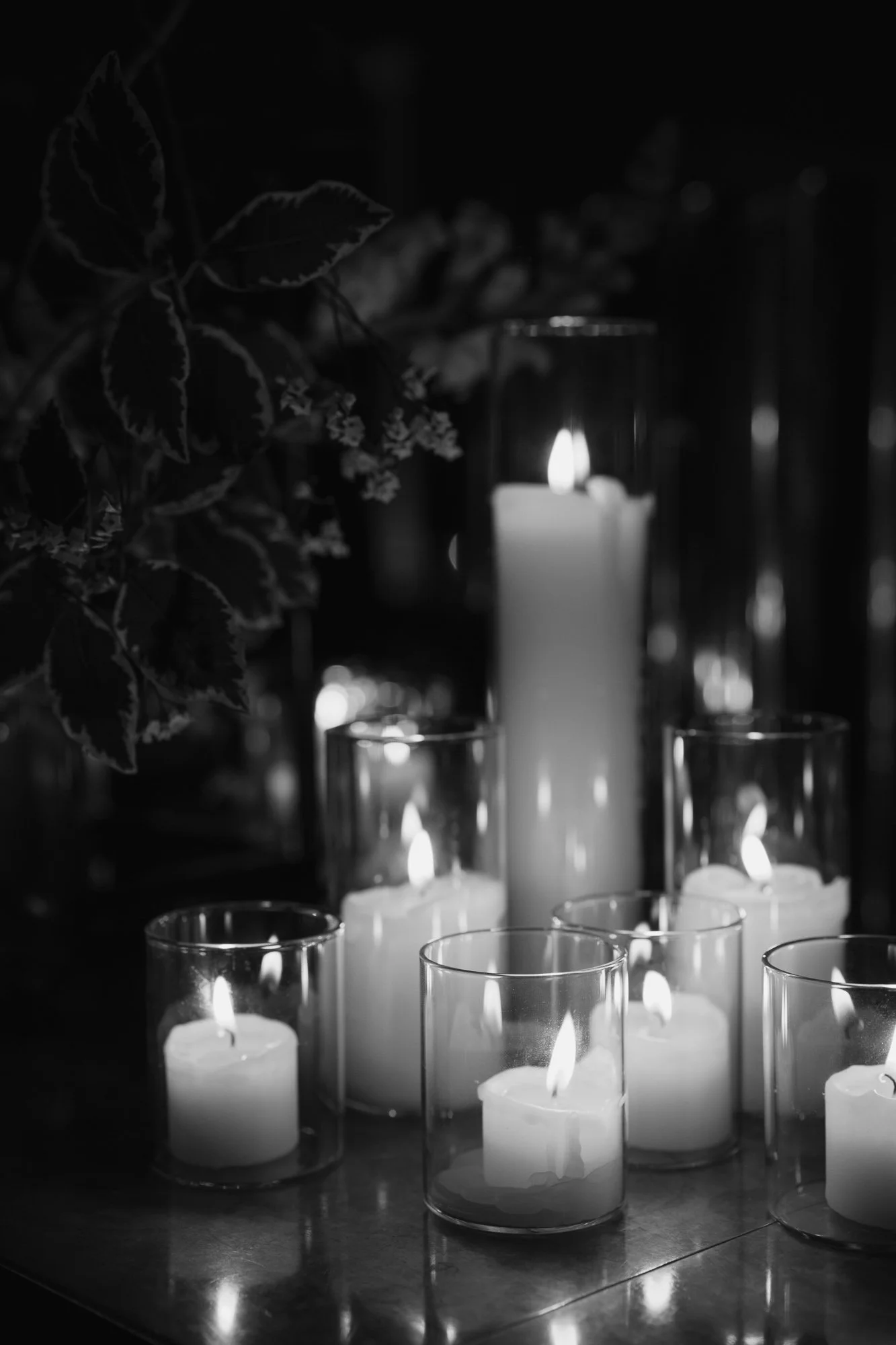 Several lit candles in glass holders, with a large candle in the background, and leaves and small flowers to the left.
