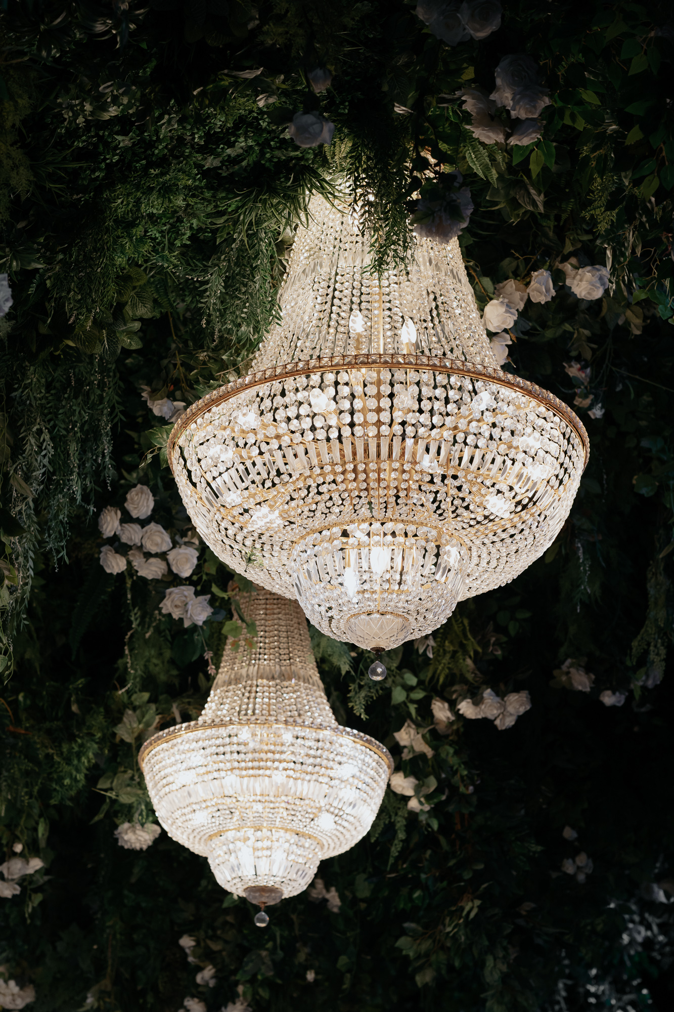 Two ornate crystal chandeliers hanging from a ceiling decorated with green foliage and white flowers at Whitestone in Bentleigh East.