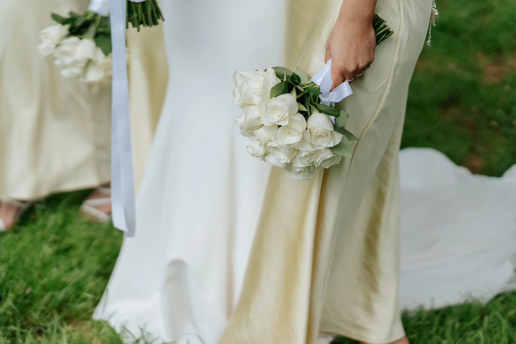 Close-up of a woman holding a bouquet of white roses at a wedding ceremony, with other bridesmaids holding similar bouquets in the background, on a grassy outdoor setting.