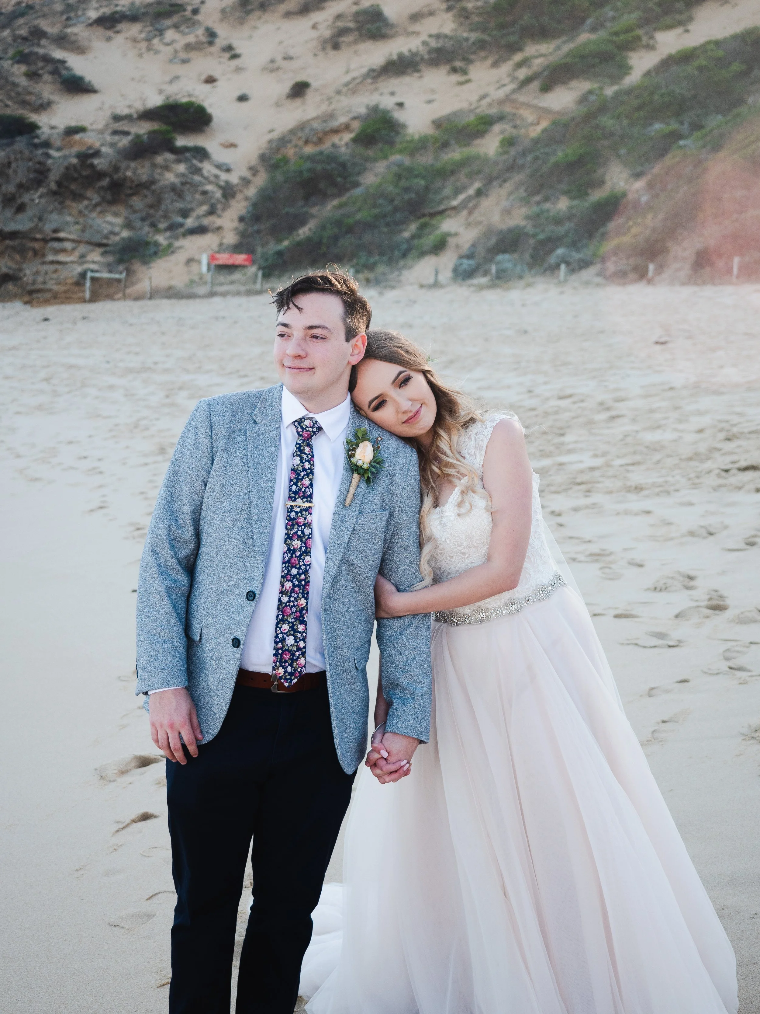 A young couple in wedding attire holding hands on a sandy beach on the Mornington Peninsula, with the woman resting her head on the man's shoulder as they pose for a photo.