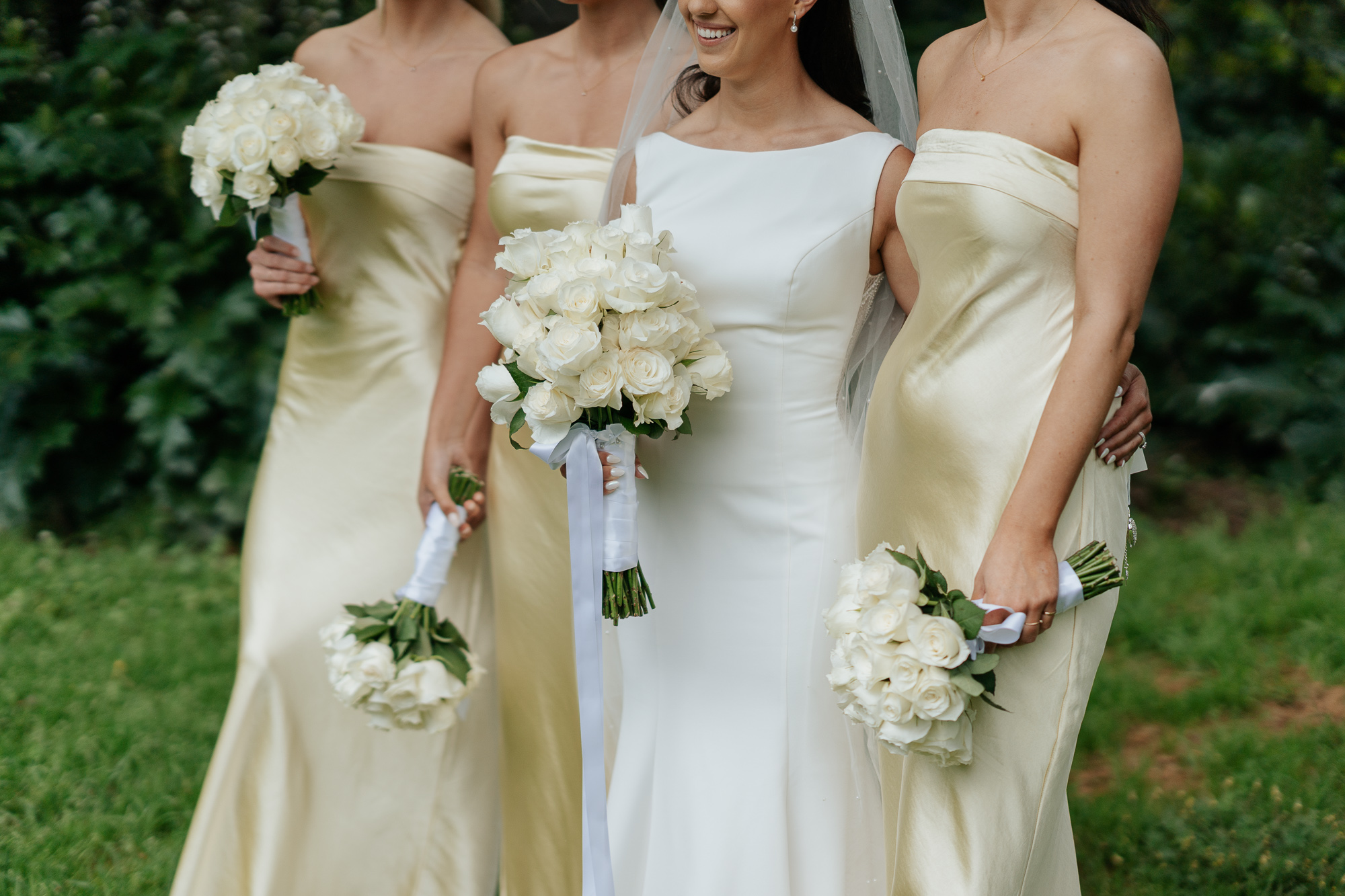 Bridesmaids in light yellow satin dresses holding white rose bouquets standing outdoors with greenery in the background. Leonda by the Yarra