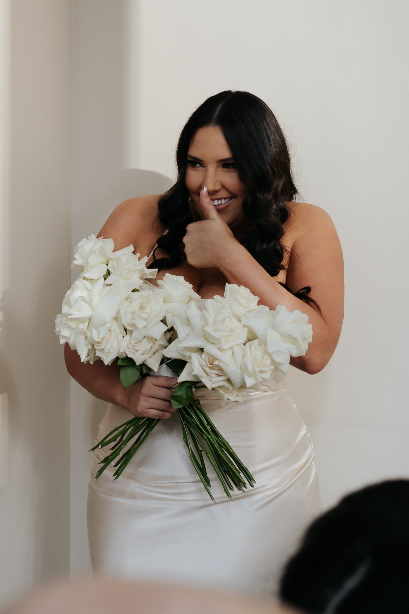 A woman with long dark hair and light skin holding a large bouquet of white roses, smiling and giving a thumbs up, wearing a satin dress.