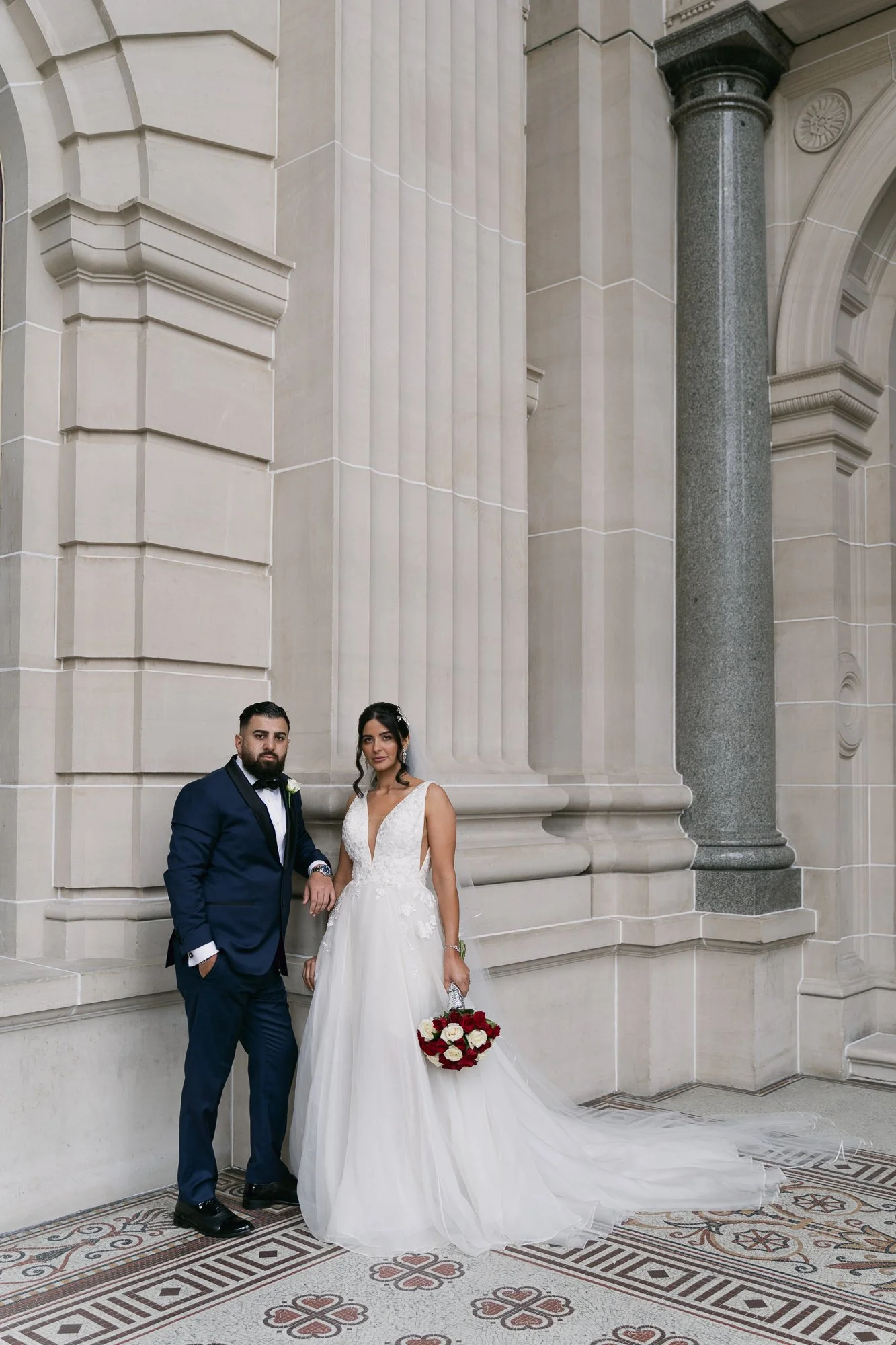 A bride and groom standing outside a grand building with large columns and ornate stonework. The bride is wearing a white wedding gown and holding a bouquet of red and white flowers, while the groom is in a navy blue suit with a white shirt and black