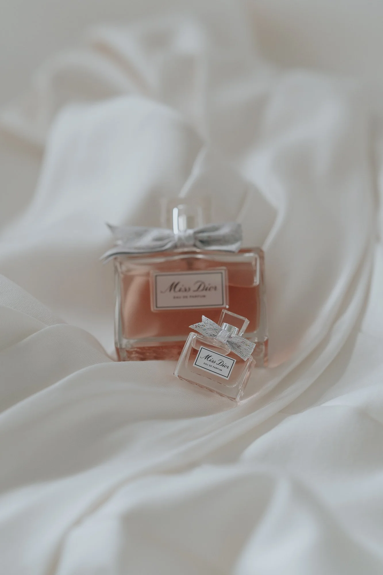Two Miss Dior perfume bottles, one large and one small, with white ribbon bows, placed on off-white fabric.