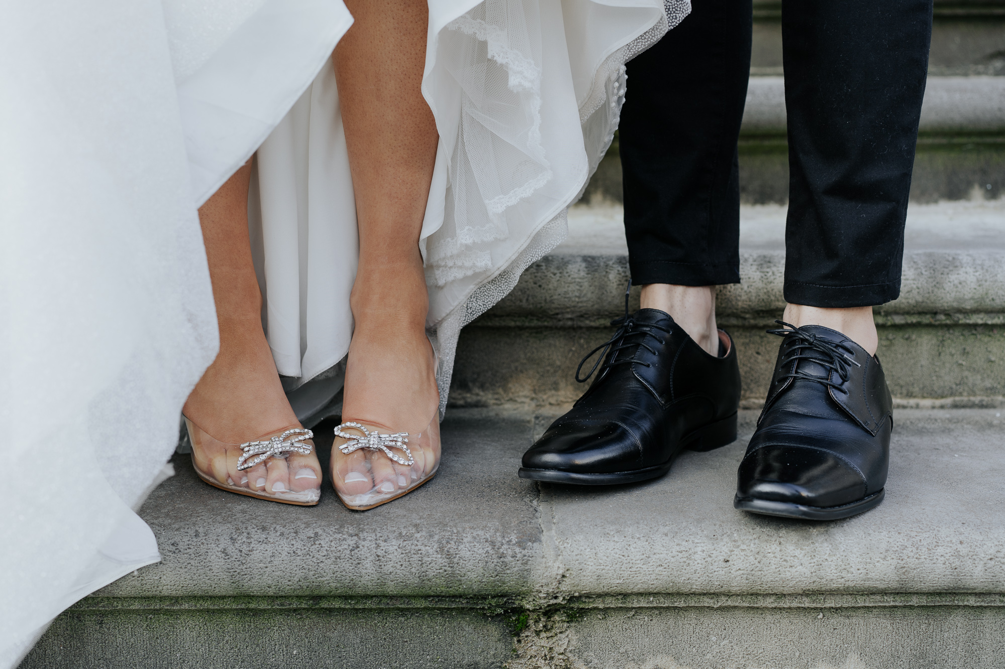 Close-up of a bride wearing elegant clear heels with bow and rhinestone accents standing on a stone step next to a groom in black dress shoes and black pants, both standing on the stone steps of Parliament House, Victoria.