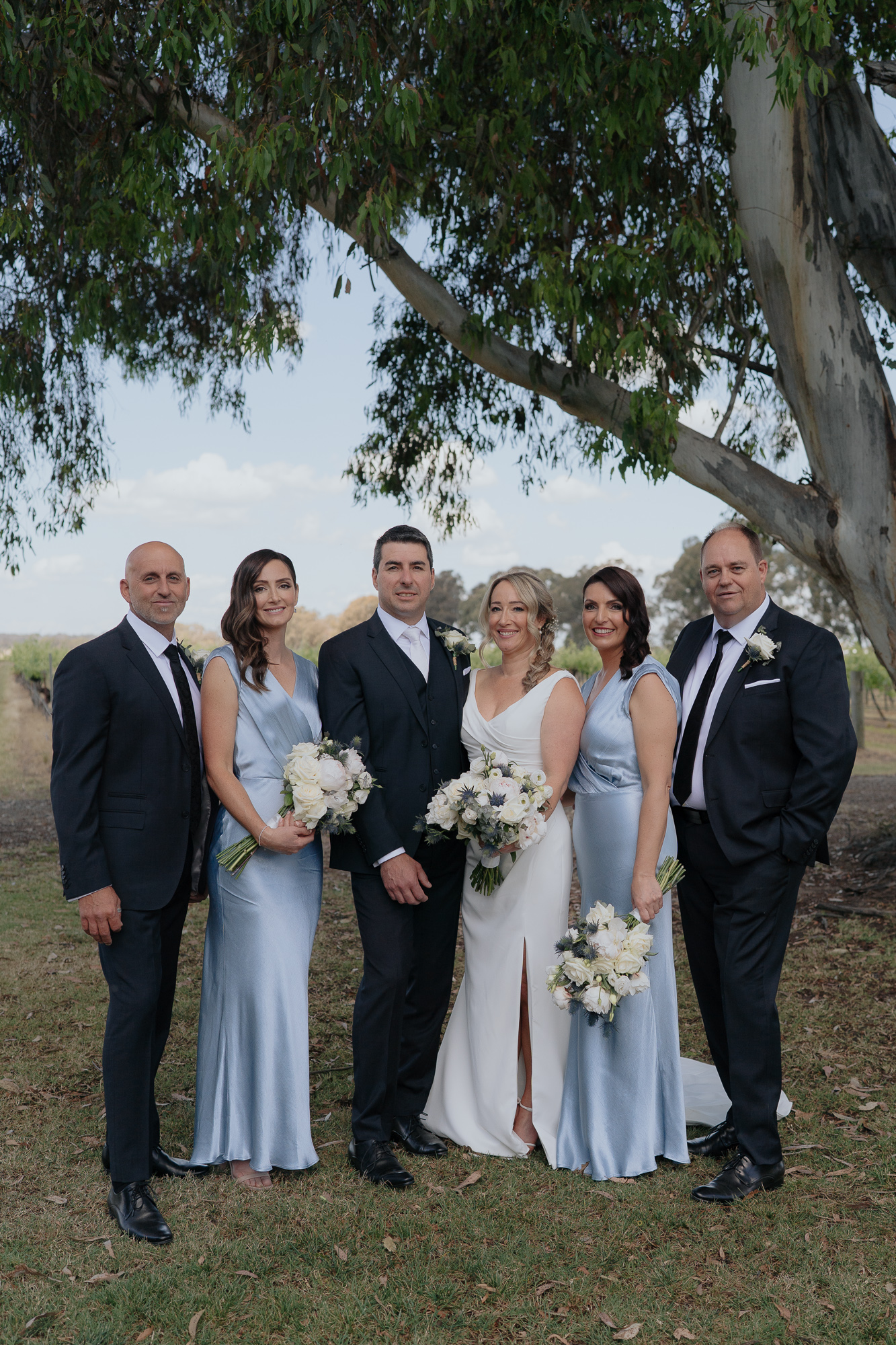 Group of six people, likely wedding party, standing outdoors under a large tree. The two women are in white and light blue dresses, holding bouquets. The four men are in dark suits, some with boutonnieres. They are smiling. Buller Wines Wedding
