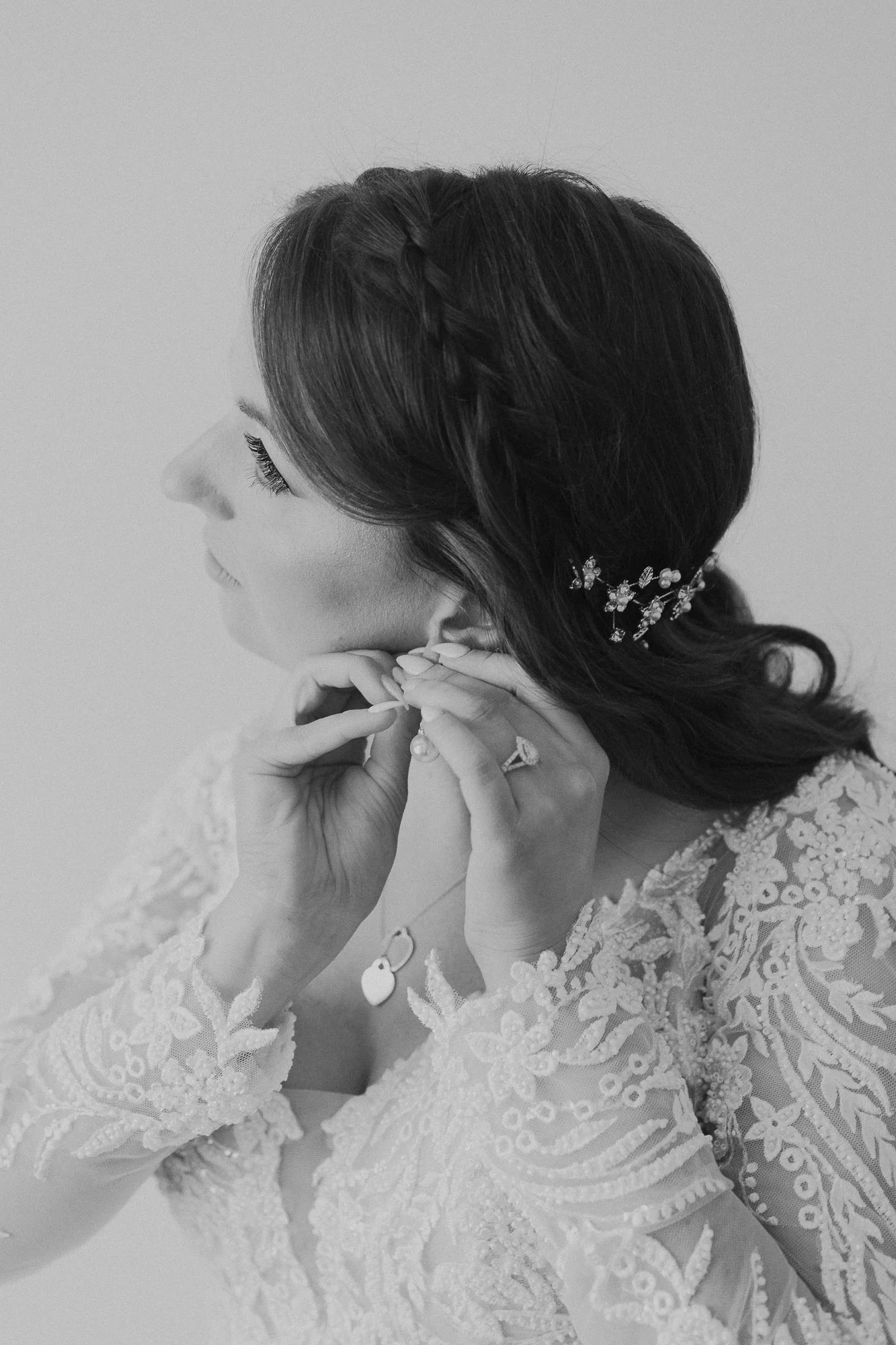 A woman with dark hair, wearing a lace dress, is putting on earrings. She has a jeweled hair accessory and is wearing a ring and necklace.