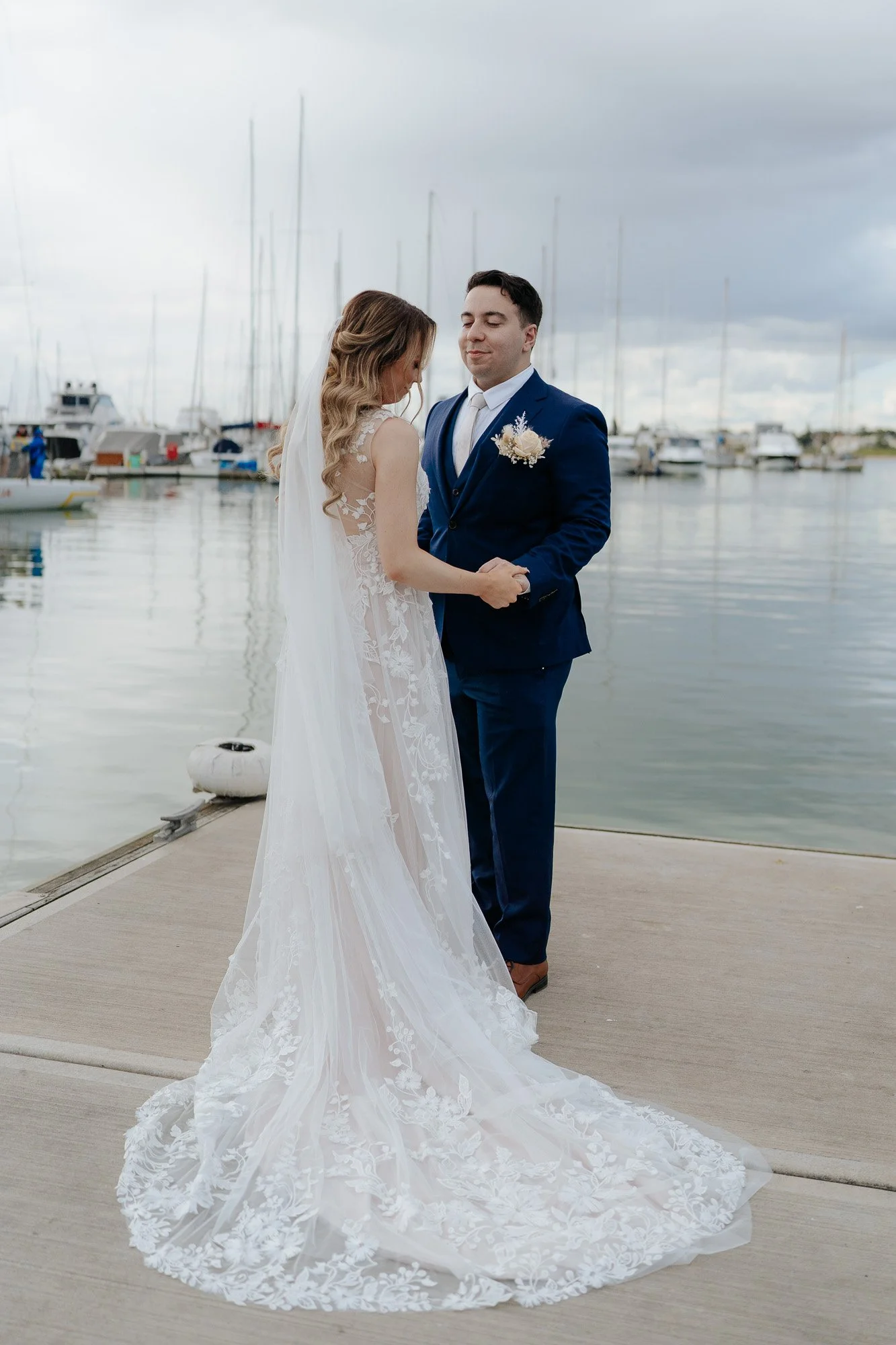 Bride and groom holding hands by the water at Sandringham yacht club, sailboats docked in the background. The bride is wearing a white lace wedding gown with a long train, and the groom is in a navy blue suit with a white shirt and boutonniere.