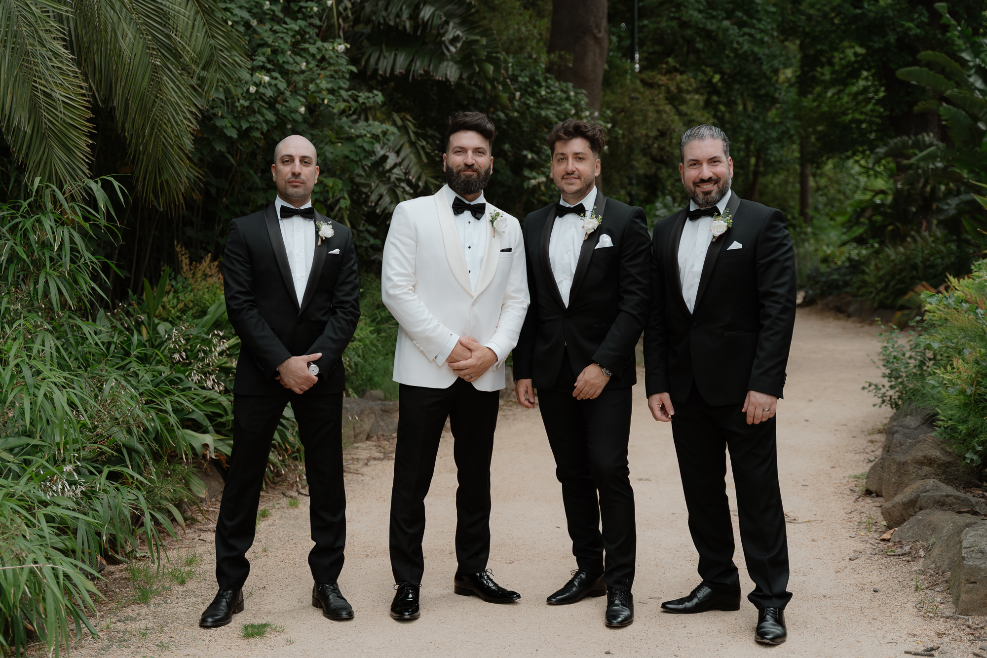 Four men in tuxedos standing on a garden path, posing for a photo at a wedding, surrounded by greenery. Fitzroy Gardens