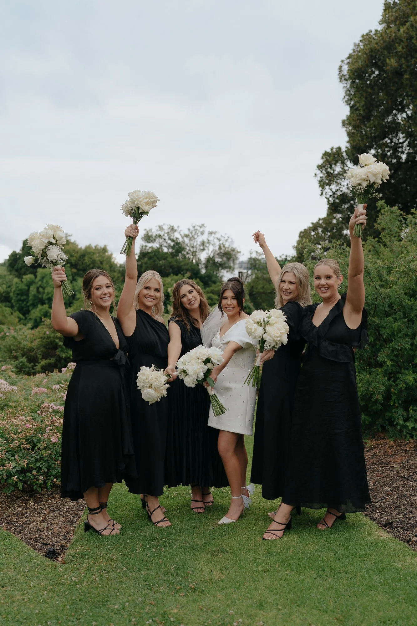 Group of six women celebrating outdoors, dressed in black dresses with the bride in white; holding bouquets of white flowers, smiling, trees and cloudy sky in background.