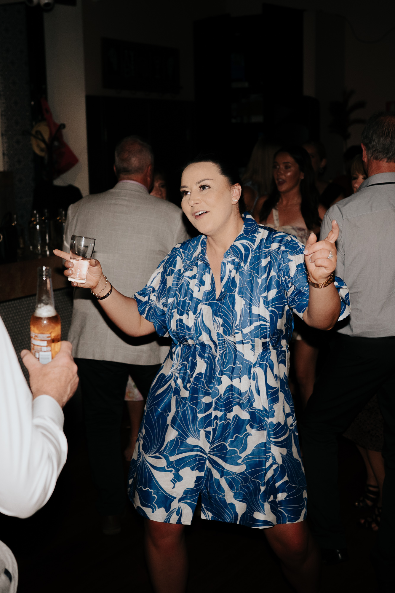 A woman in a blue and white floral dress dancing at a party with drinks in her hands, surrounded by other guests.