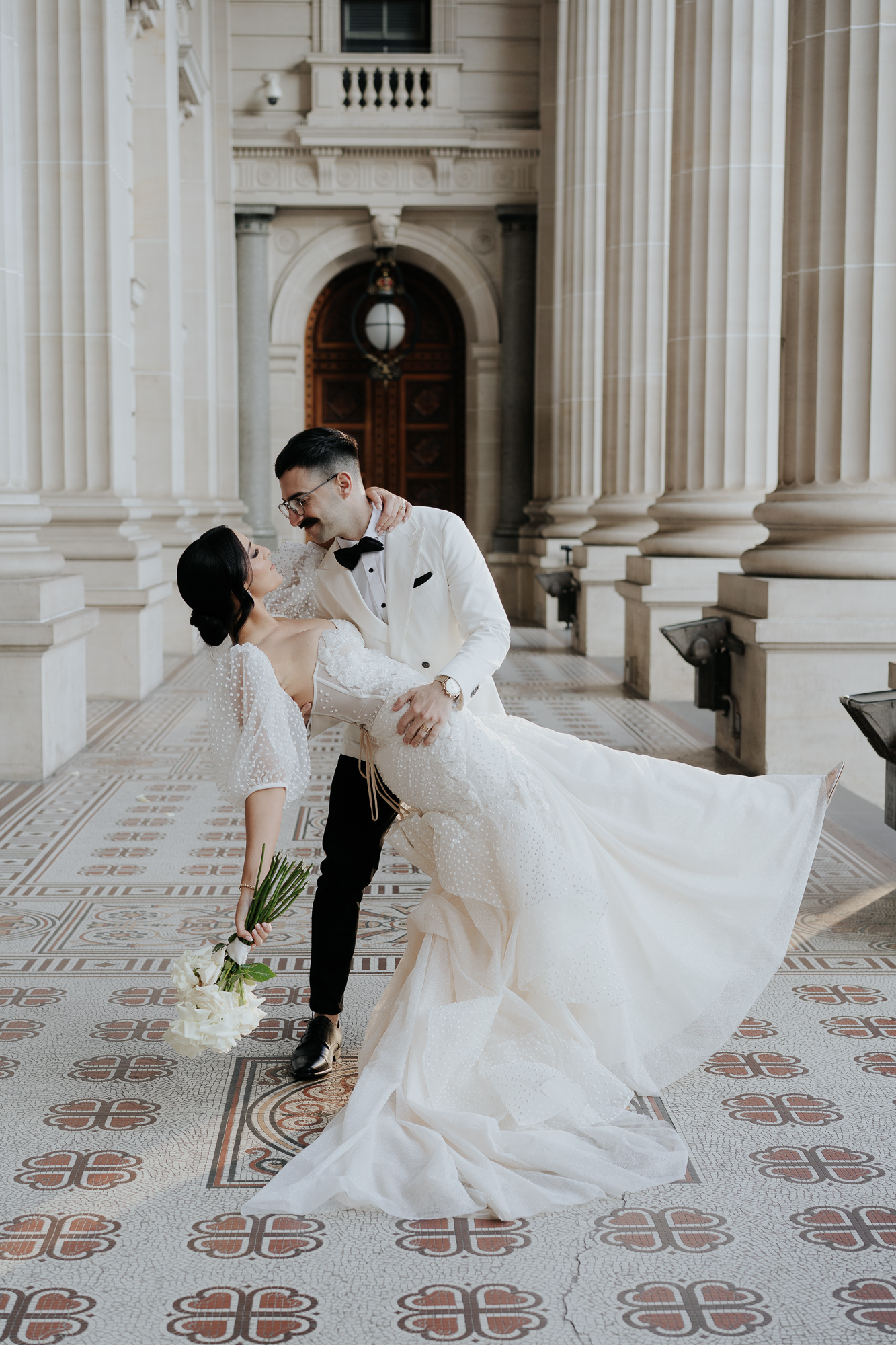 A bride and groom in elegant wedding attire at Parliament House Melbourne. The groom, in a white tuxedo, dips the bride in a romantic pose, holding a bouquet of white flowers. The bride wears a white wedding gown with puffed sleeves.