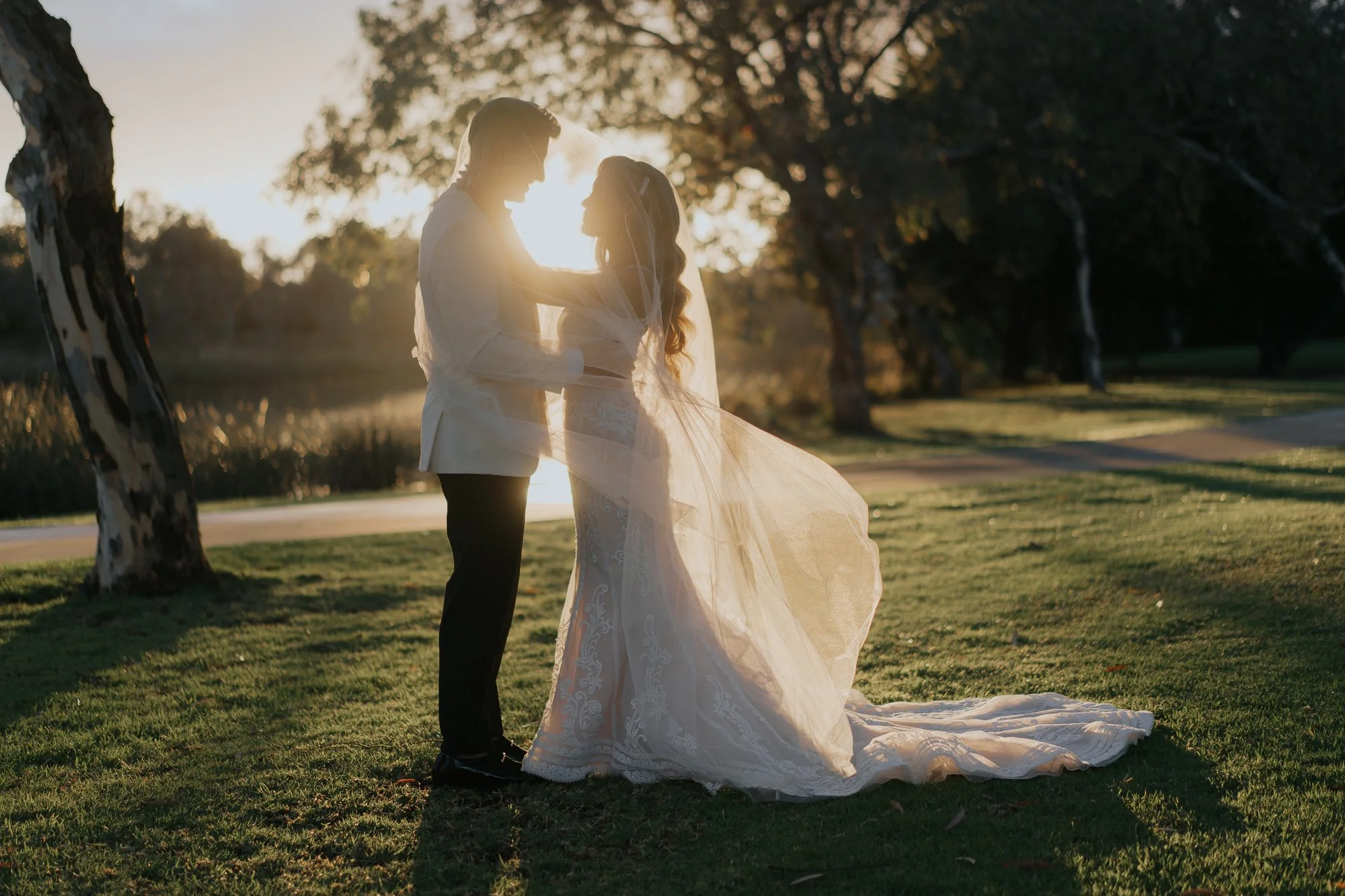 A bride and groom are standing close together outdoors during sunset, with the groom gently holding the bride's face. The bride is wearing a white lace wedding gown and veil, and the groom is in a white suit jacket and dark pants. They are surrounded