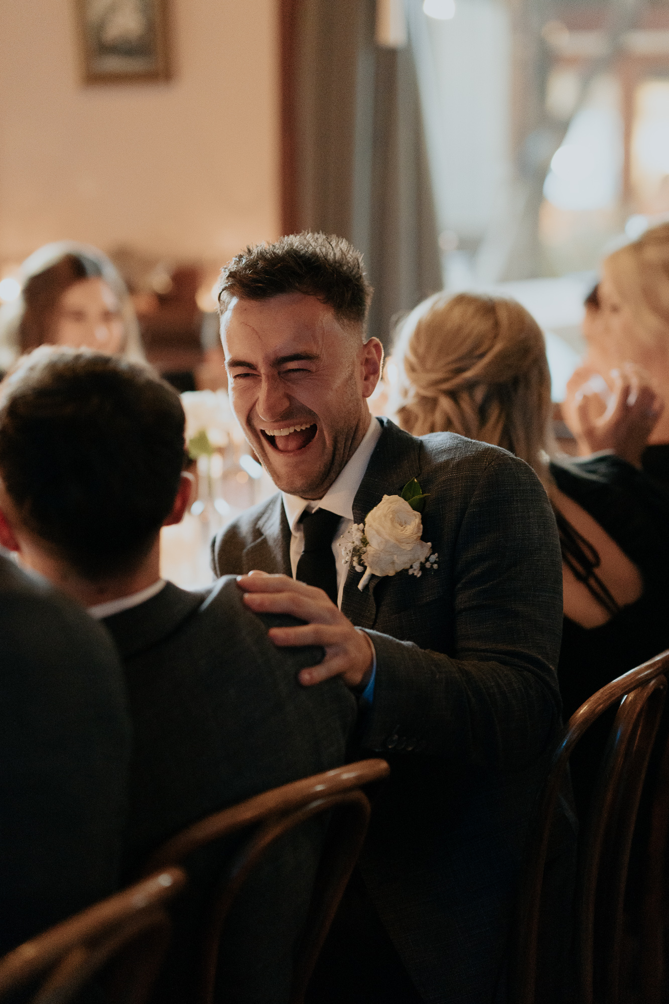 A man in a suit and tie laughing and engaging in conversation at a wedding or formal gathering, with other guests seated around him.