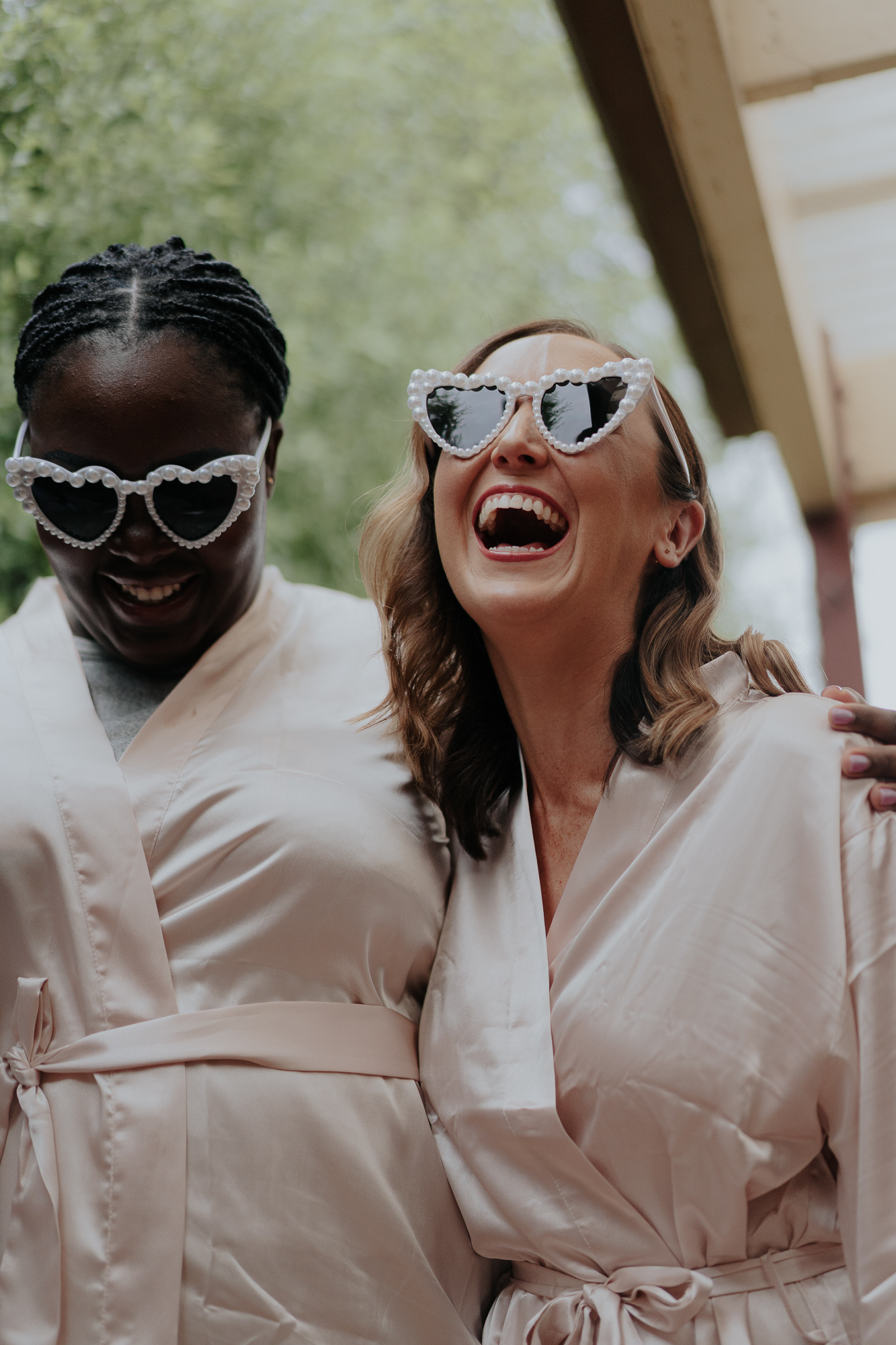 Two bridesmaids wearing matching satin robes and sunglasses, laughing and sharing a joyful moment outdoors.