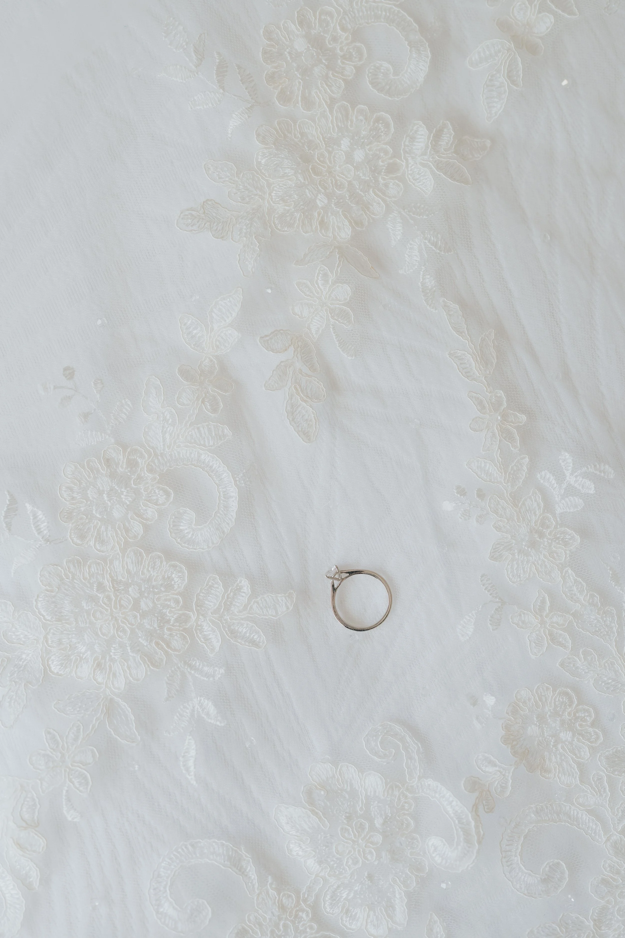 A close-up of a white, embroidered fabric with floral patterns and a silver ring placed on it.