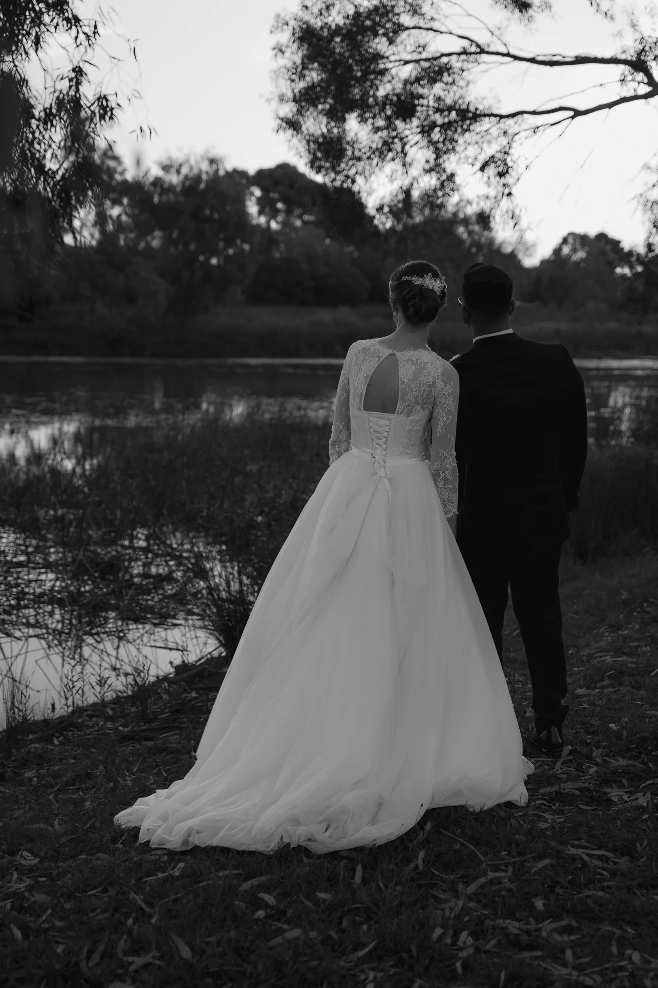 A bride and groom standing by a river at sunset, with the bride wearing a long, lace wedding gown and the groom in a black suit. At the Lakeside Banquet and Convention Centre.
