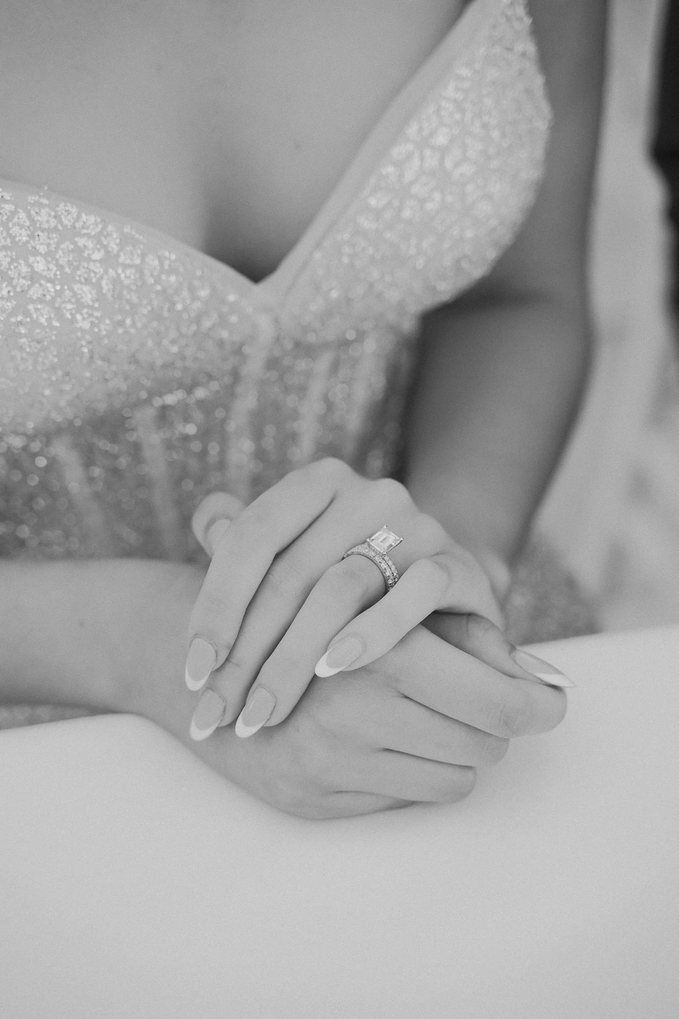 Close-up of a woman's hands with an engagement ring and wedding band, resting on a white surface, wearing a glittery dress.