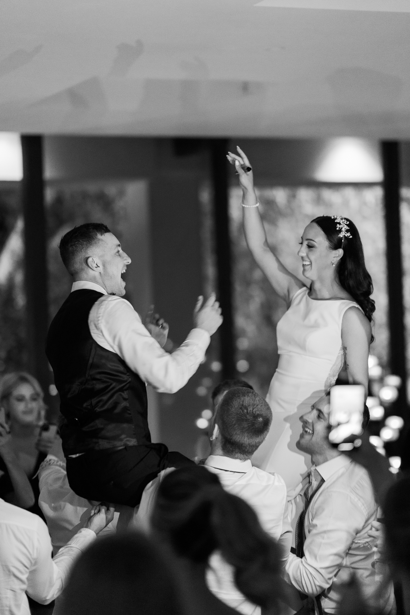 A bride and groom dancing joyfully at their wedding reception, with the groom seated on a chair and the bride standing, surrounded by guests, in a black and white photo.