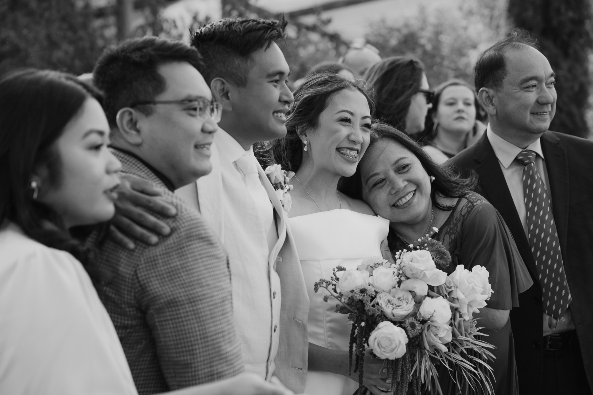 Black and white photo of a diverse group of people at a wedding, smiling, with the bride holding a bouquet of flowers.