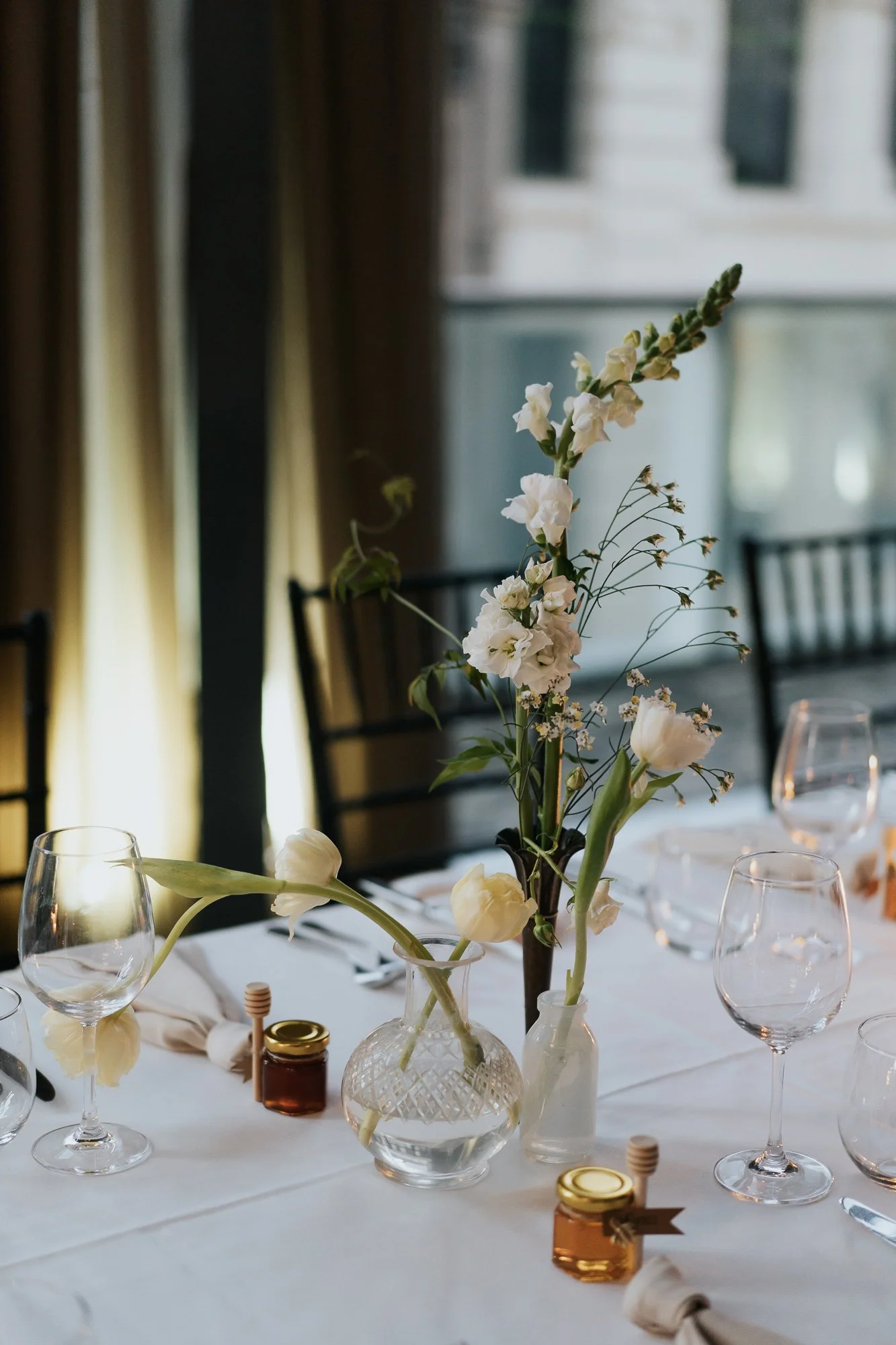 Elegant table centerpiece featuring white flowers in clear vases on a white tablecloth, surrounded by wine glasses, small jars, and napkins, set in a softly lit room with dark curtains and window views.
