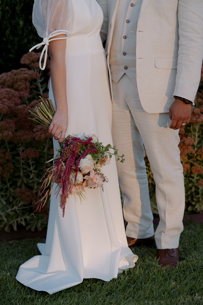 A bride and groom standing outdoors at Olivehouse at Greendale Grove, with the bride holding a bouquet of flowers, both dressed in wedding attire.