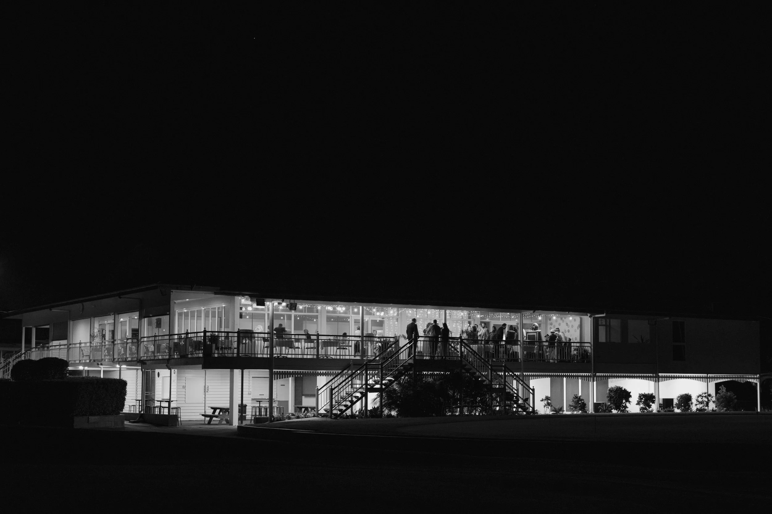 Night view of Wynnum Golf Club with illuminated interior and people inside, seen from set of stairs outside.