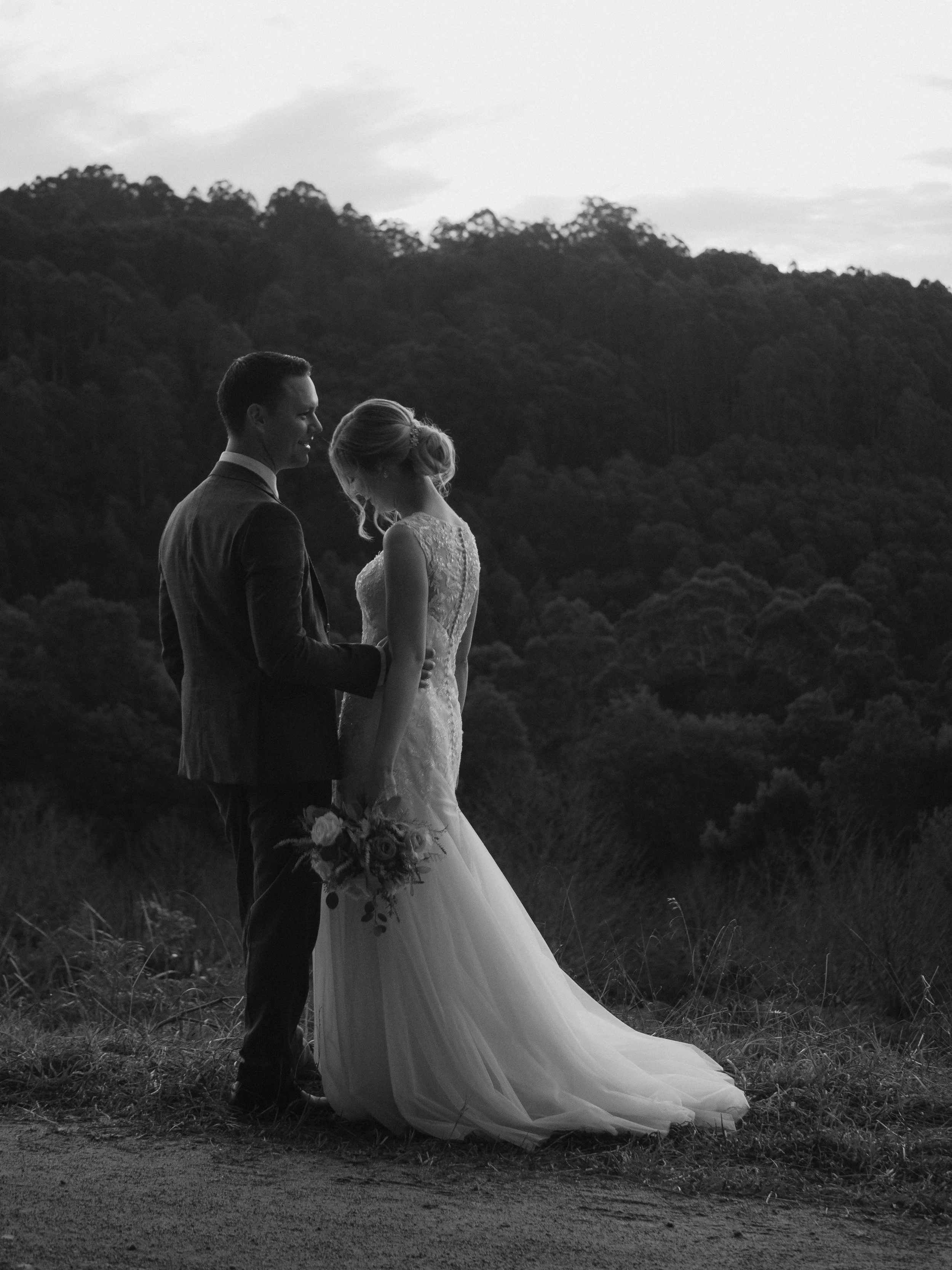 Black and white photo of a bride and groom standing close outdoors, with the groom holding the bride's hand and smiling, she is holding a bouquet, and they are in front of a forested hillside.