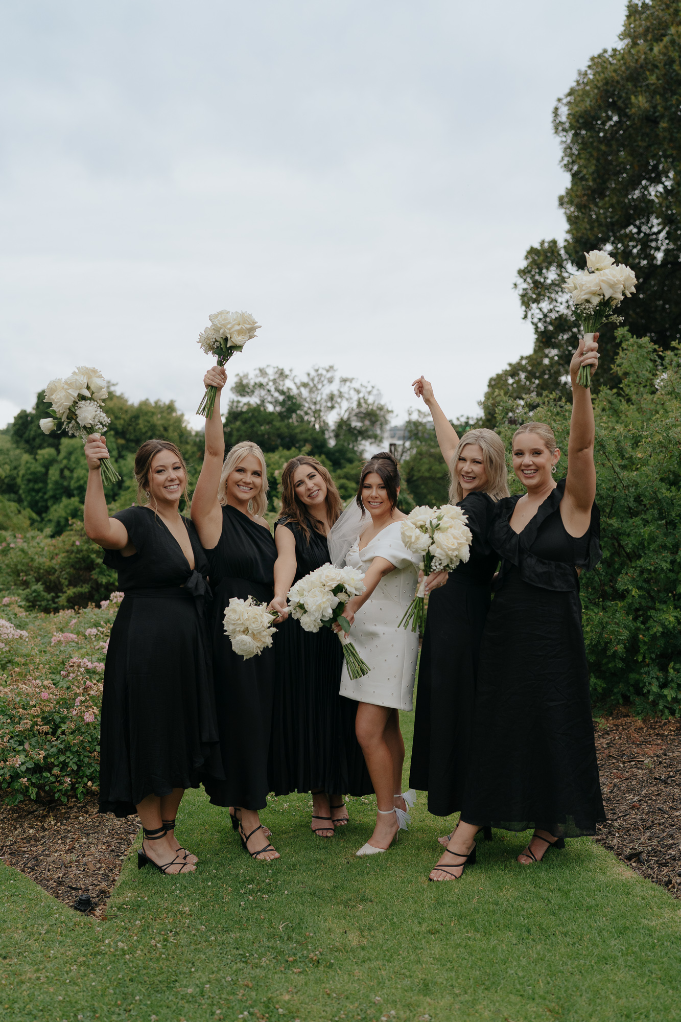 A group of six smiling women dressed in black and white dresses, holding white bouquets, celebrating outdoors in a garden setting. Royal Botanical Gardens Melbourne