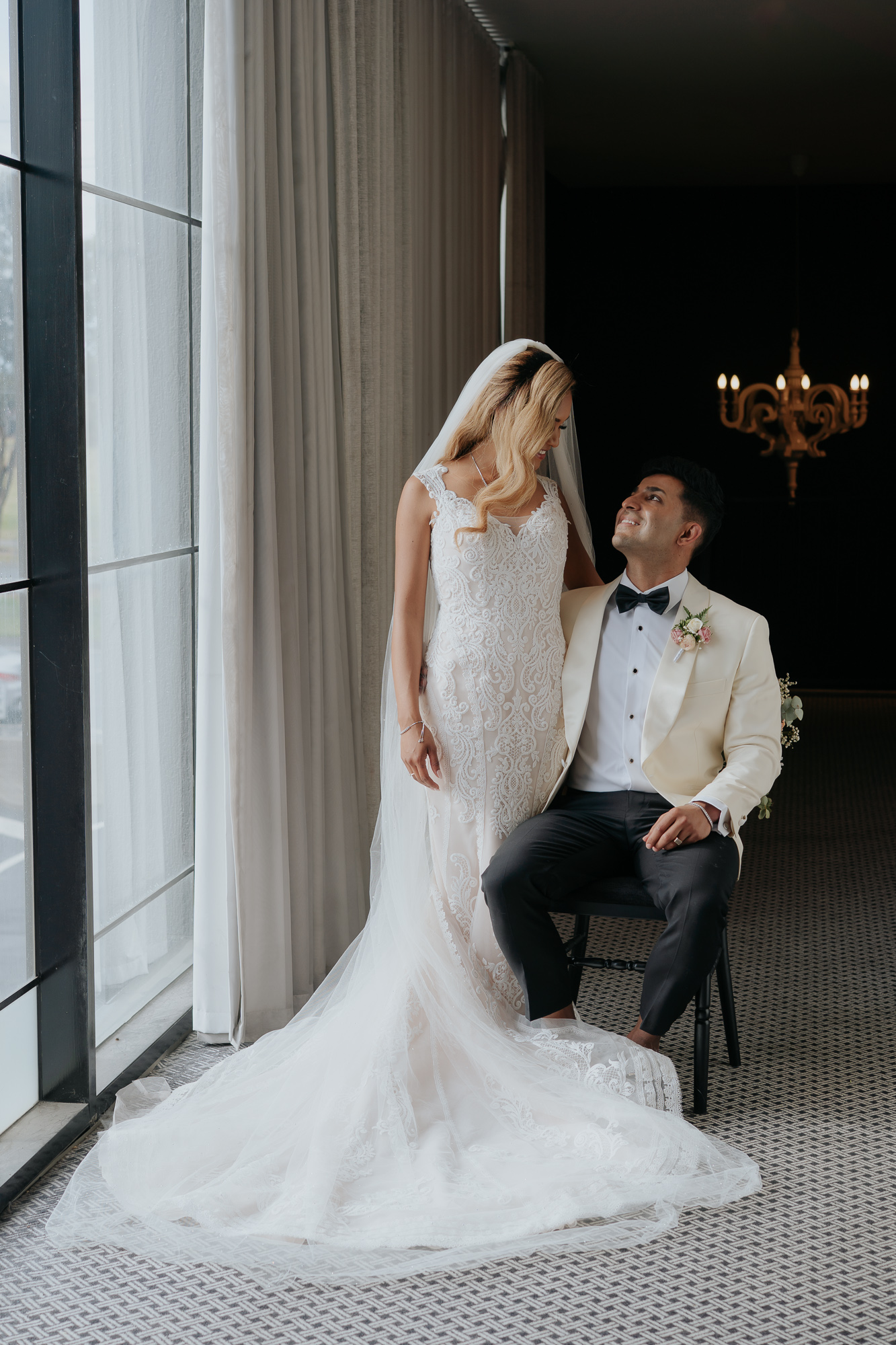 A bride and groom inside a room, with the bride standing in a white wedding gown and the groom sitting on a black chair wearing a white tuxedo jacket with a black bow tie, looking at each other affectionately. Lakeside Events