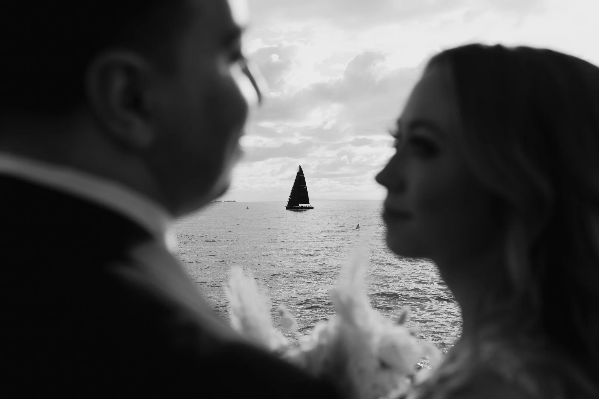 Black and white photo of a couple facing each other with a sailboat in the background on the water at Sandringham Yacht Club.