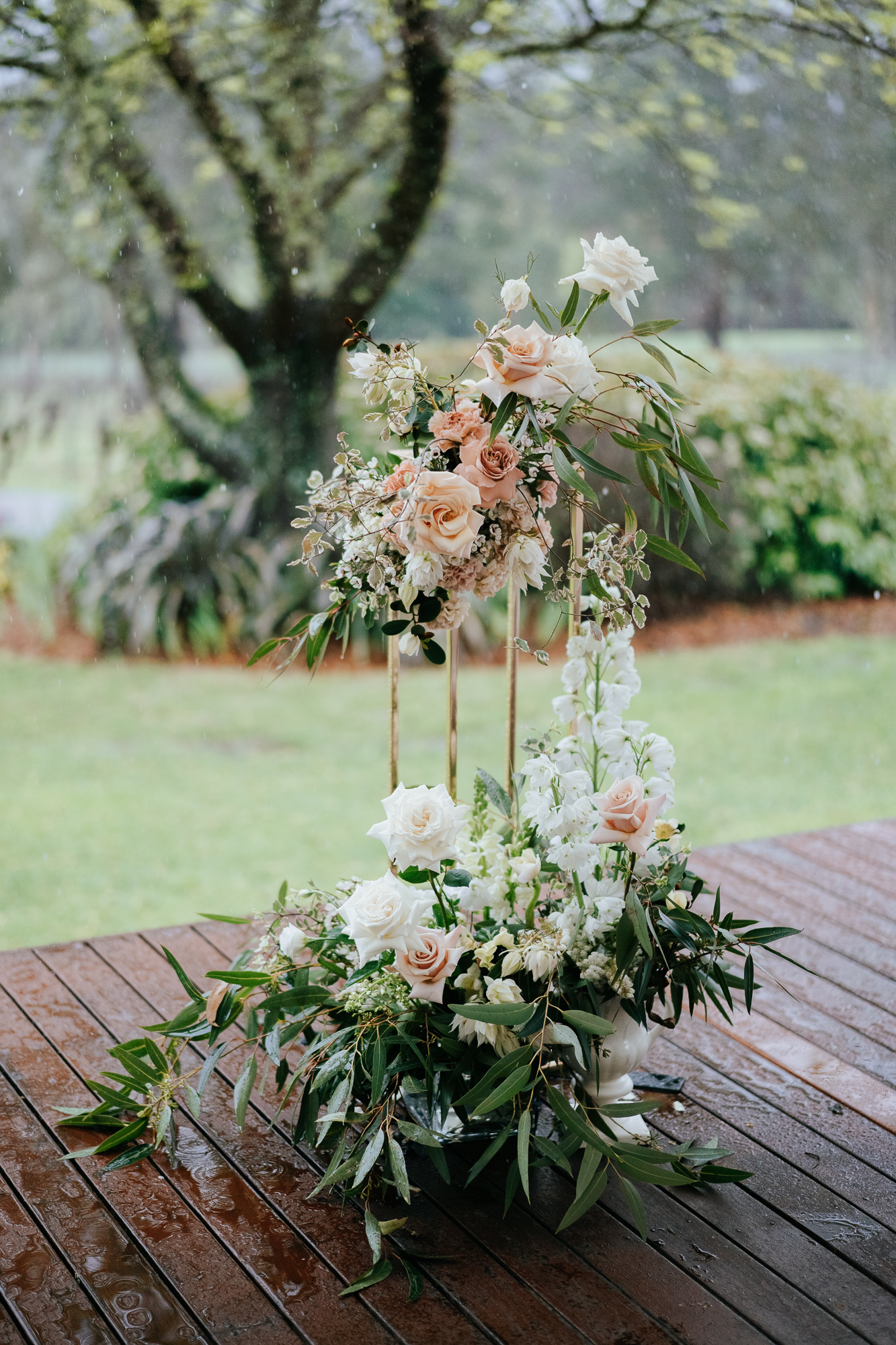Flower arrangement with white and blush roses, white delphiniums, and greenery on a wooden table, with a rainy outdoor background.