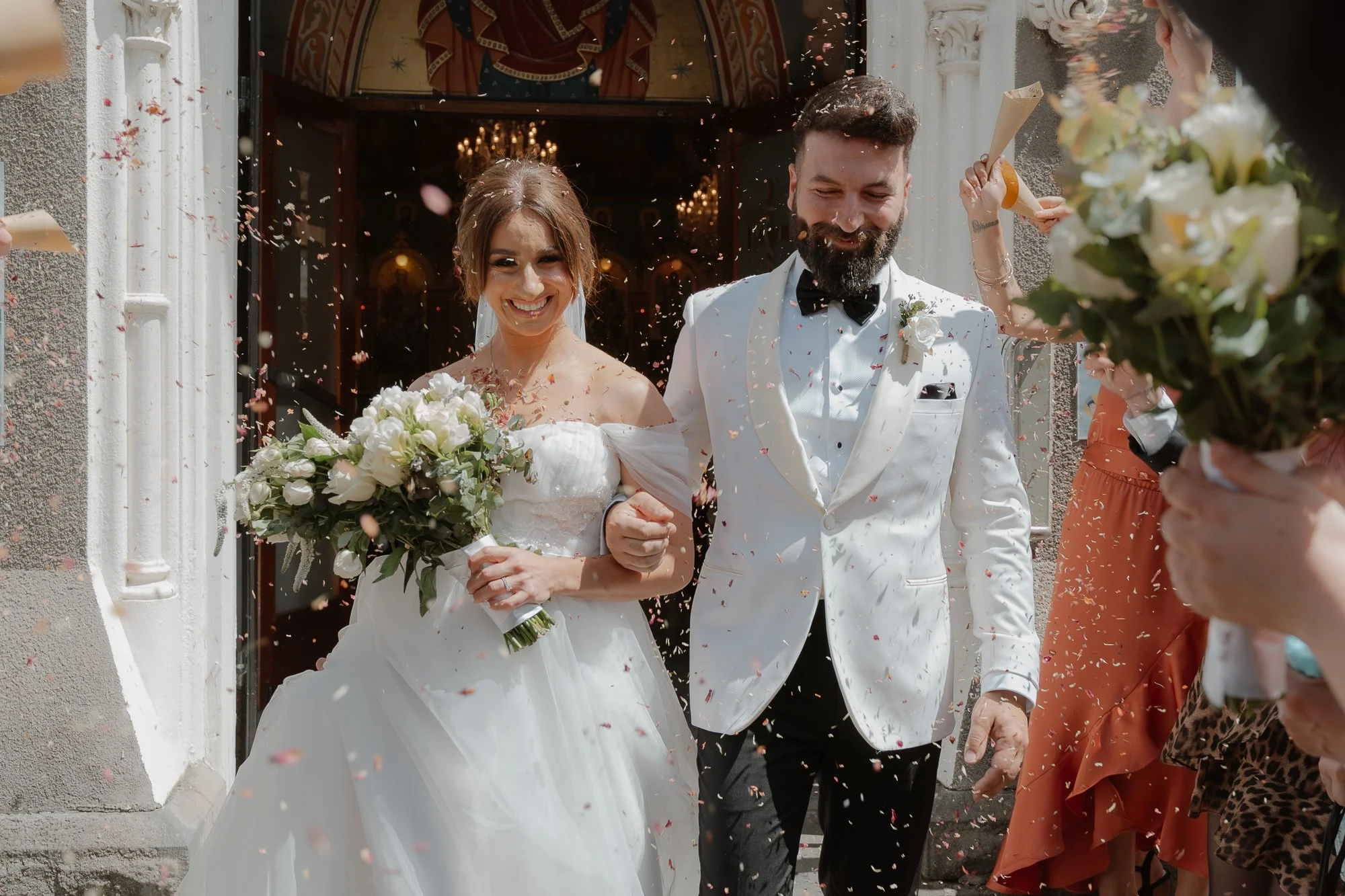Happy newlywed couple walking out of a church, celebrating their wedding with guests throwing confetti. The bride is holding a bouquet and wearing a white wedding dress, the groom in a white tuxedo jacket with a black bow tie.