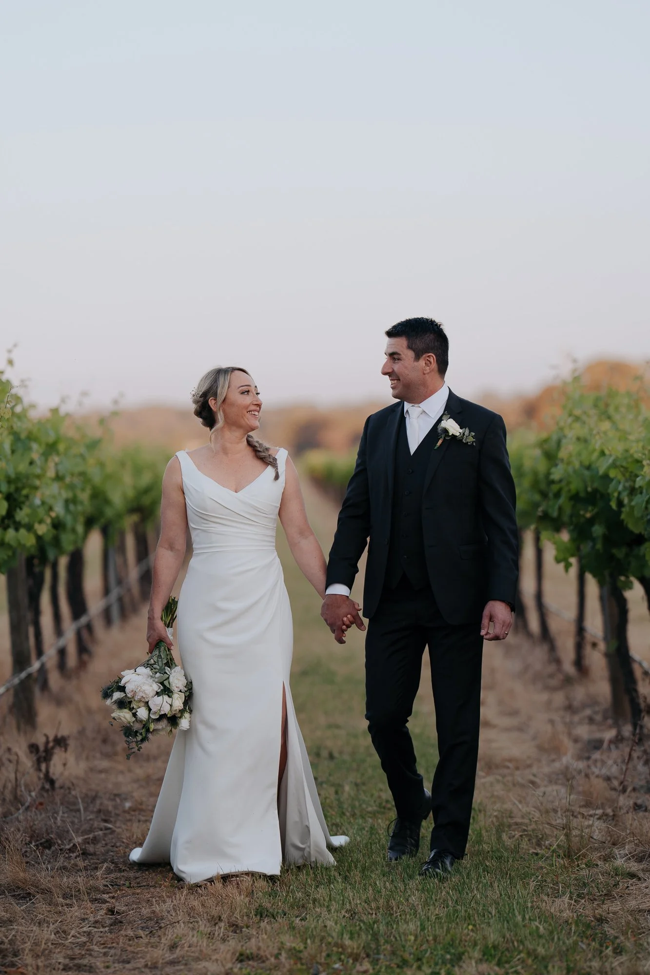 A bride and groom walking hand in hand through a vineyard at Buller Wines Rutherglen, smiling at each other. The bride is holding a bouquet of flowers, and the groom is dressed in a black suit. It is sunset.