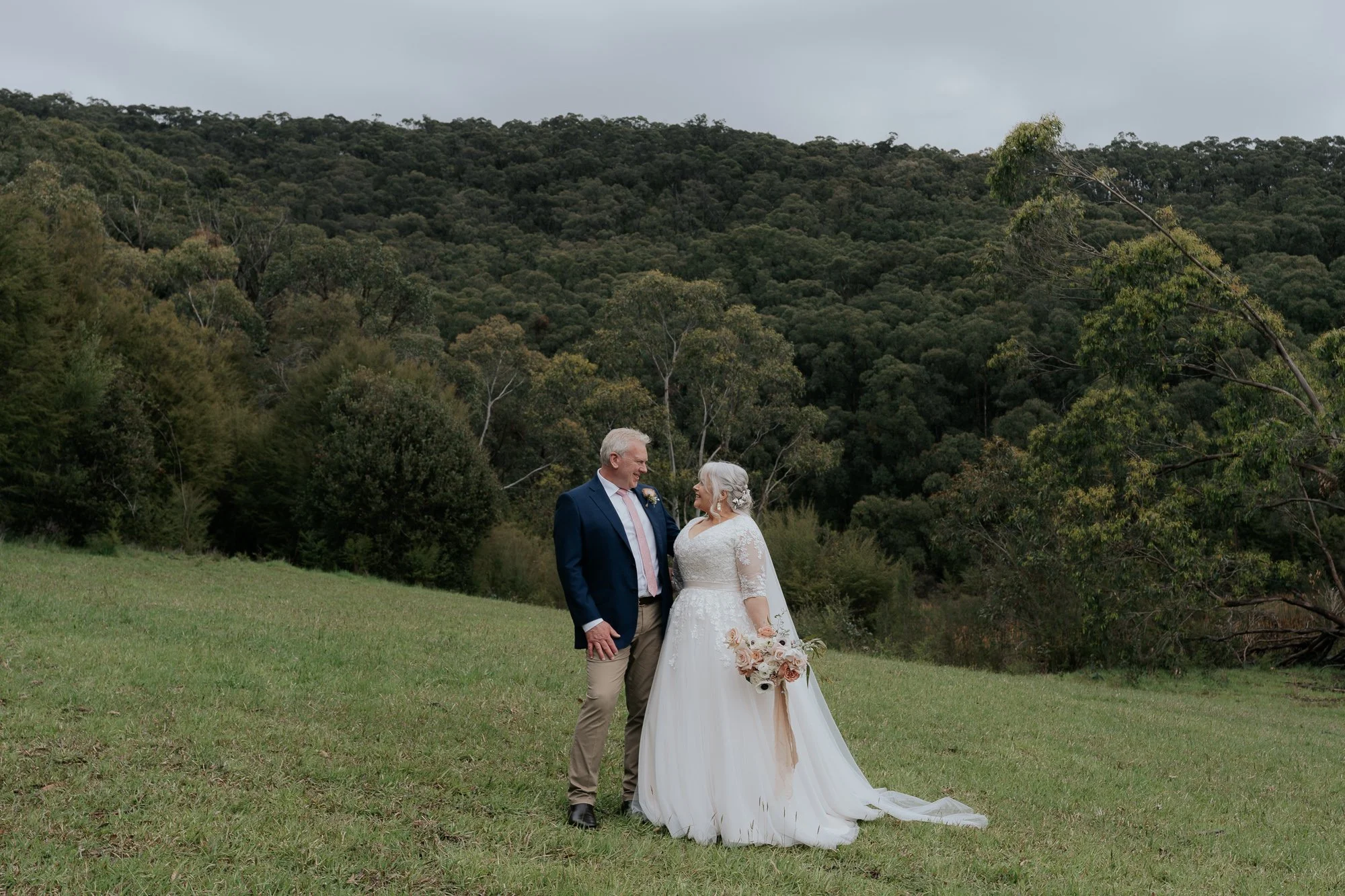 Bride and groom standing in a grassy field with trees and hills in the background, smiling at each other after wedding.